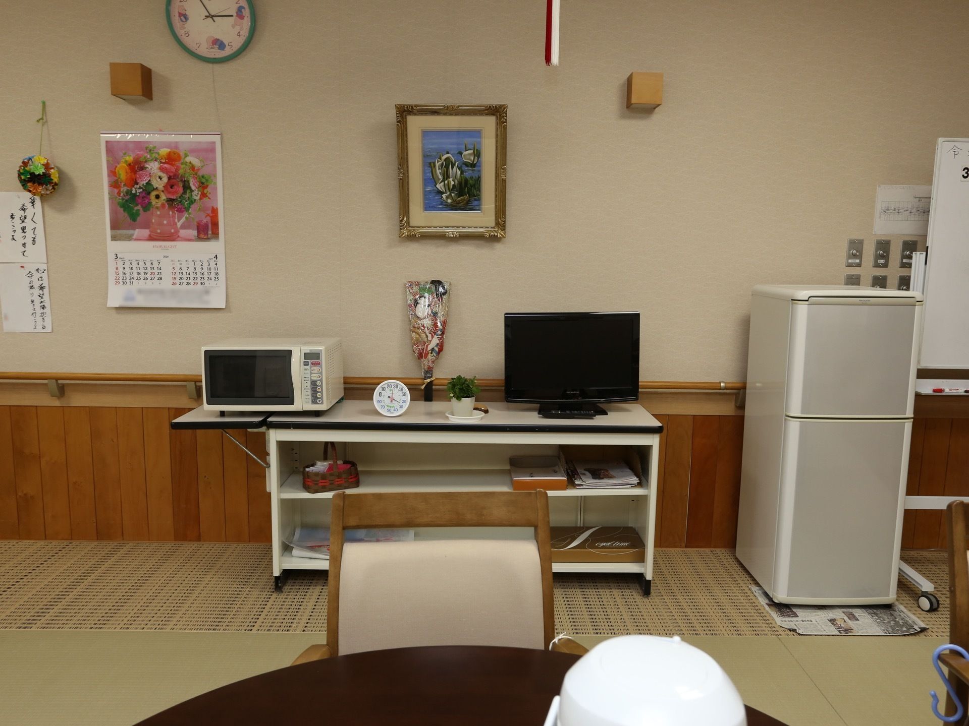 手すり付きの白い壁際には、冷蔵庫が配置されている。横には可動板付きの白い収納棚が置かれ、上には時計やテレビ、電子レンジが配備されている。