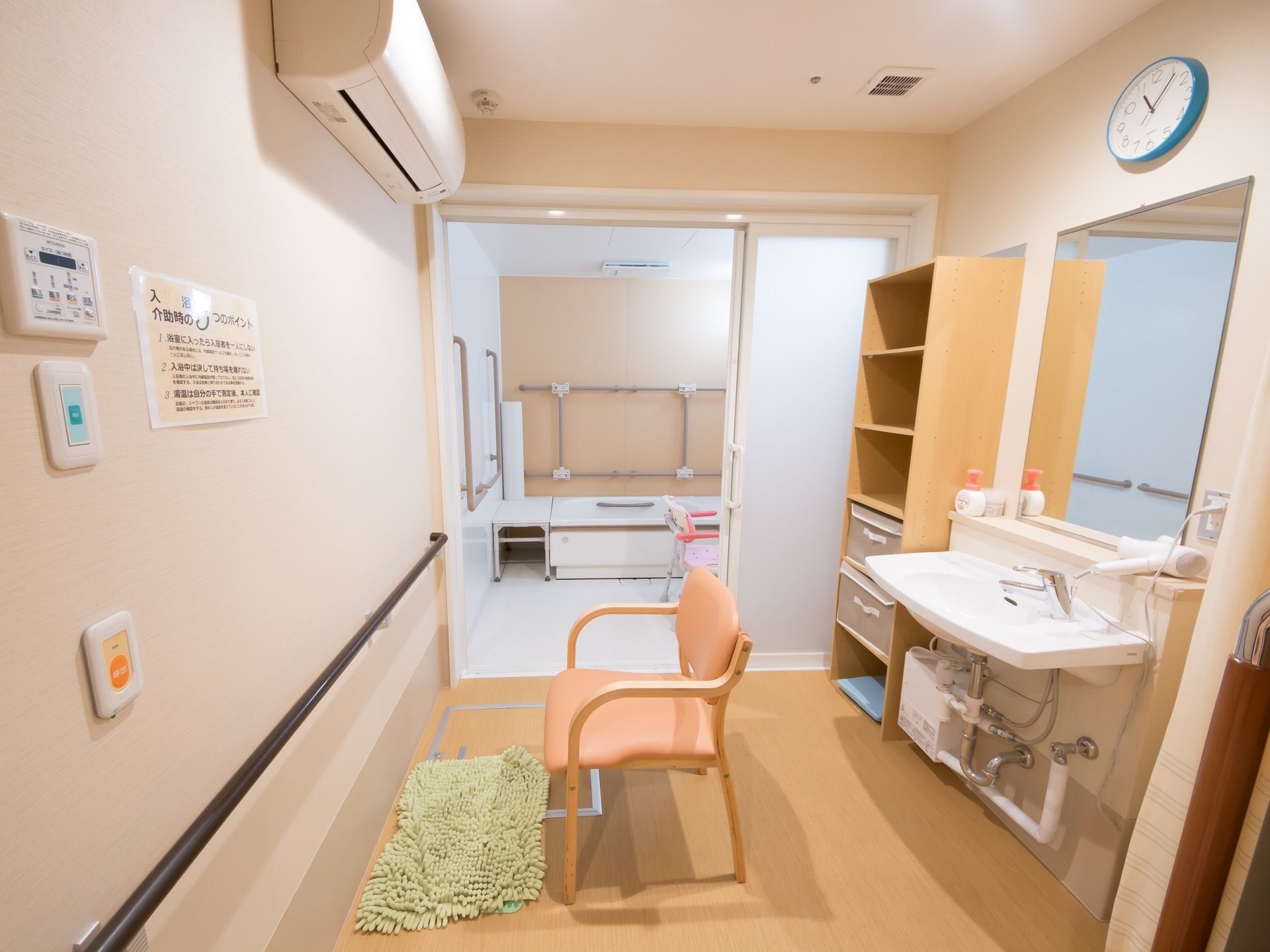 浴室の脇の着替え室にはエアコンの他に鏡付きの洗面台、衣類を置く棚、椅子、マット、ナースコールが備わっている。壁には手すりが設置されている。