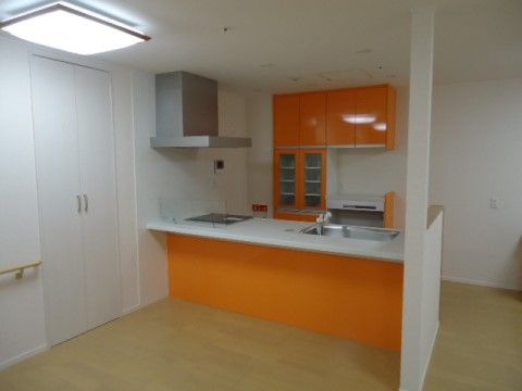 キッチンは対面式で背面に収納棚が設置されており、白い壁にオレンジ色が映えている。カウンター周りはスッキリと片付けられている。