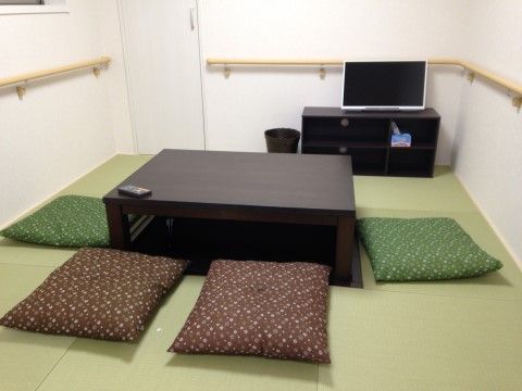 室内の中央に濃い茶色の掘りごたつがあり、周囲に緑や茶色の座布団が並んでいる。テレビは低い場所に置いており、壁には手すりが設置されている。