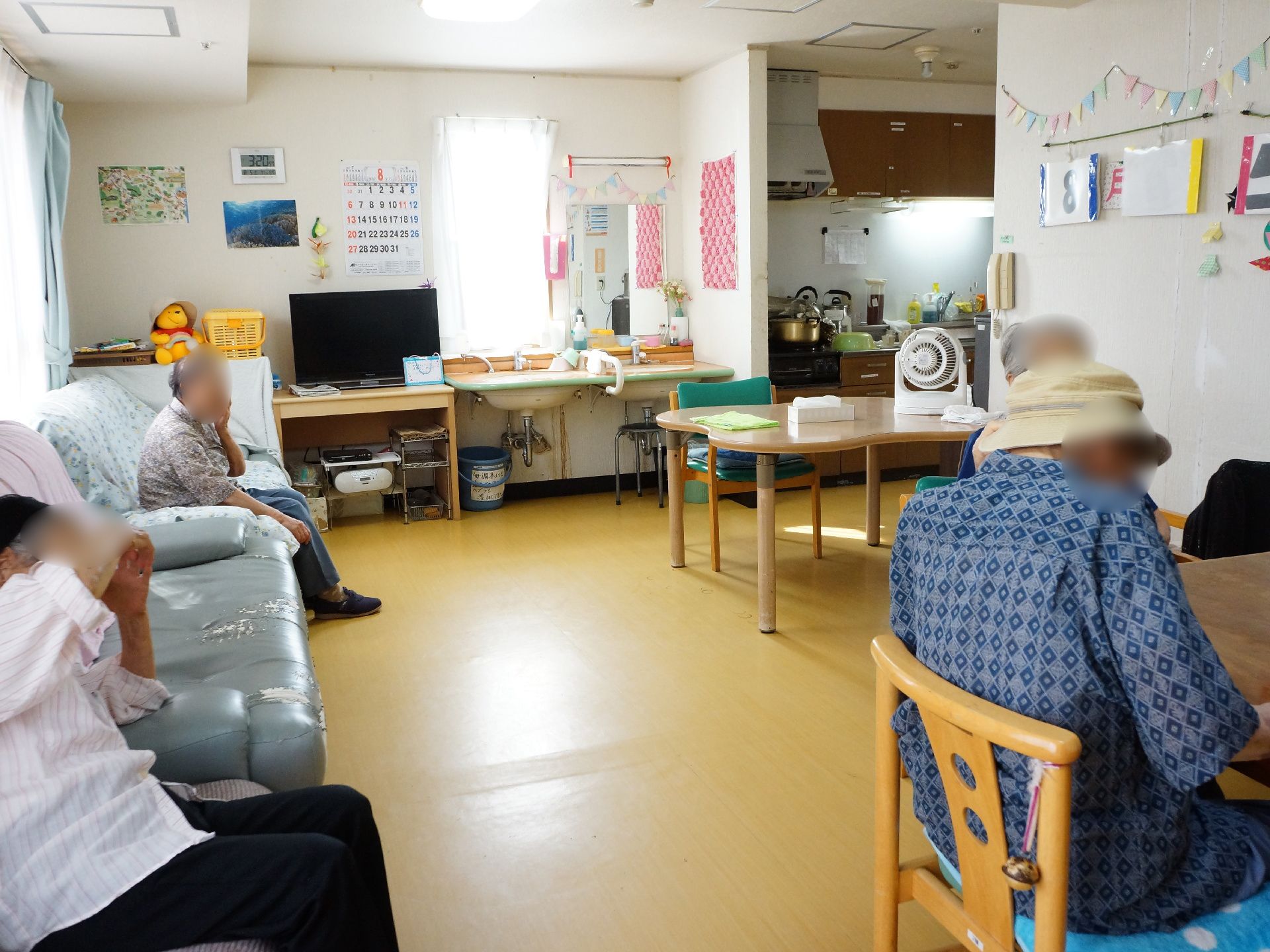 エアコンが完備の部屋には、ソファが数台、テーブル、いすなどが設置してある。手を洗うための洗面台がある。入居者が安らいでいる。