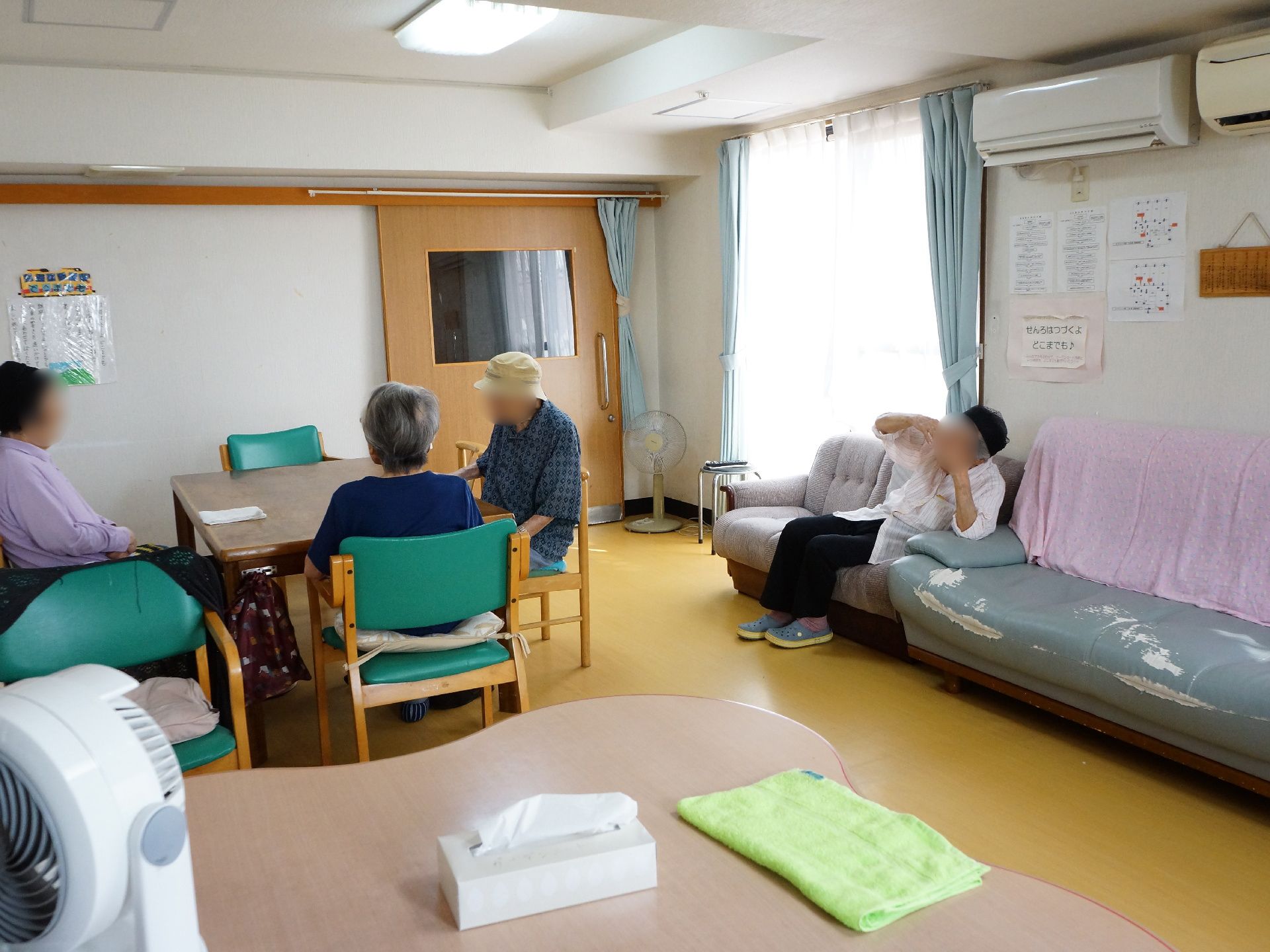 ソファ、椅子とテーブルが置いてある部屋で、入居者が椅子に座って話したりしている。壁には、絵と歌の歌詞が貼ってあり、娯楽の場である。