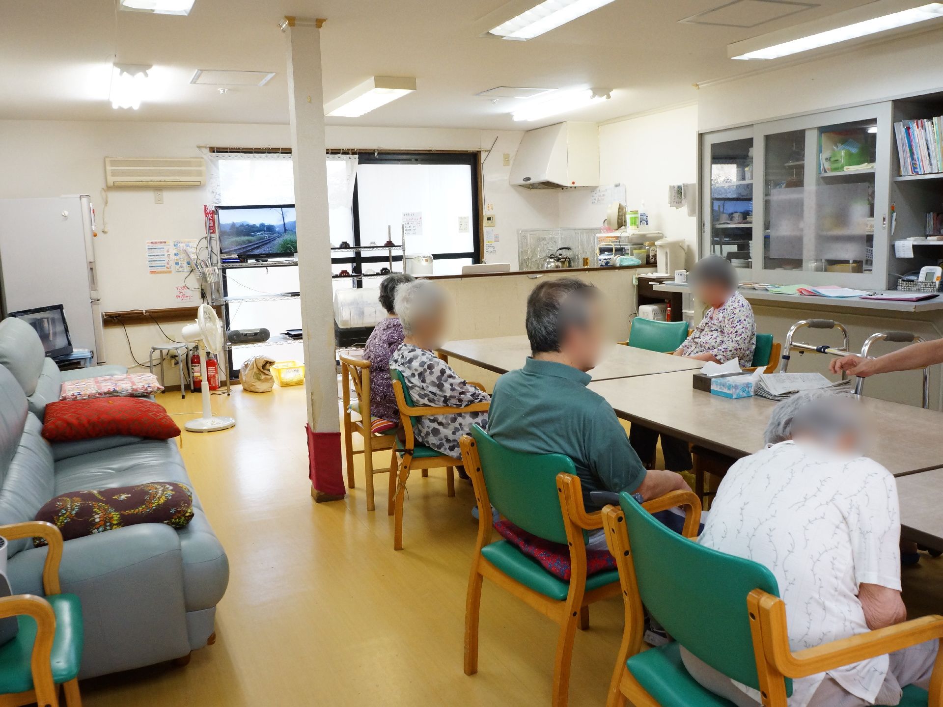 コンパクトなキッチンがついた食堂で、机が数台並べてあり、入居者が肘付きの椅子に腰かけている。テレビ、ソファがあり、憩いの場でもある。