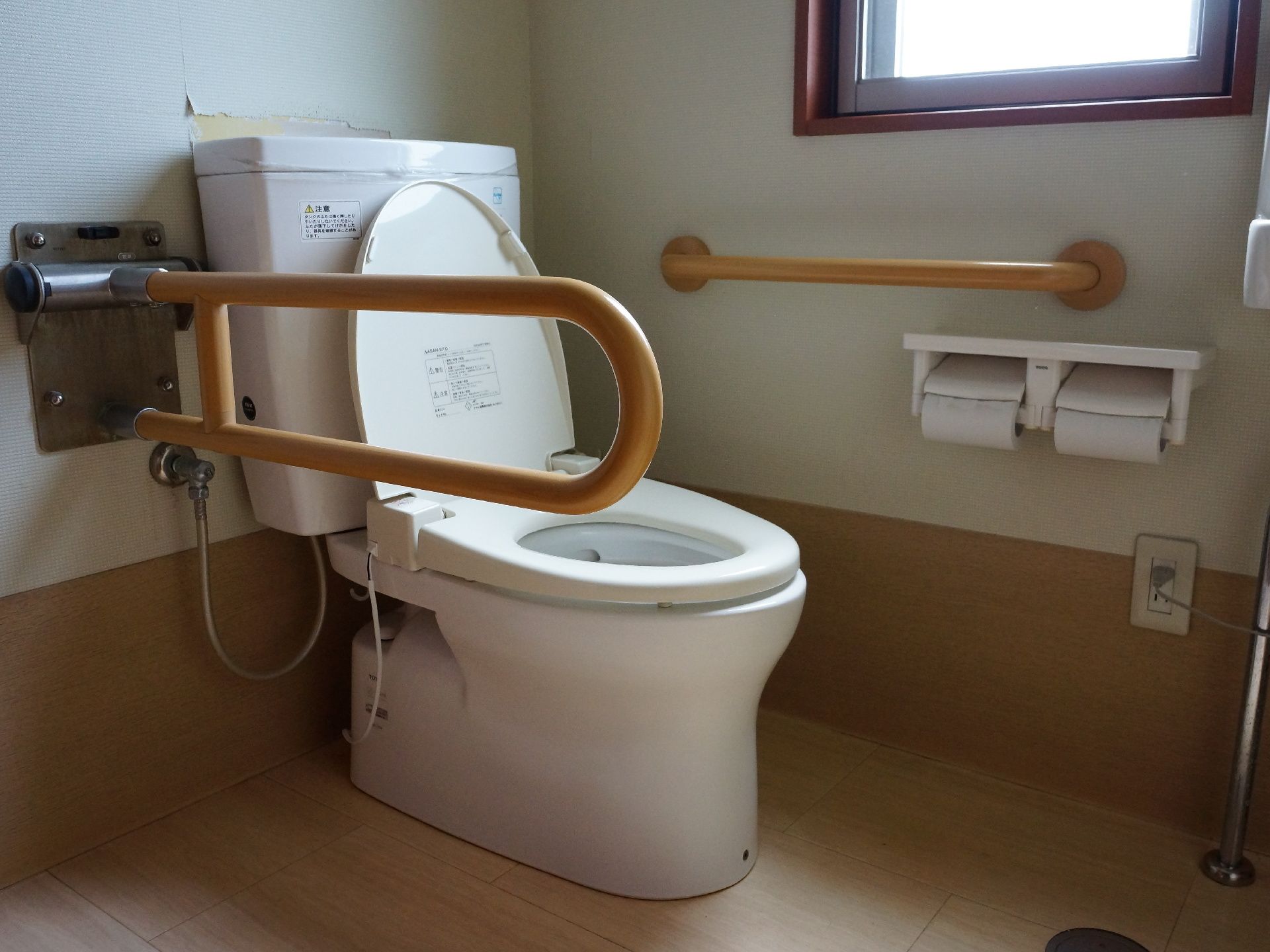 フラットなフロアにトイレが設置されている。洋式便器の左右には手すりがあり、左側の壁には窓が設けられている。コンセントがある。
