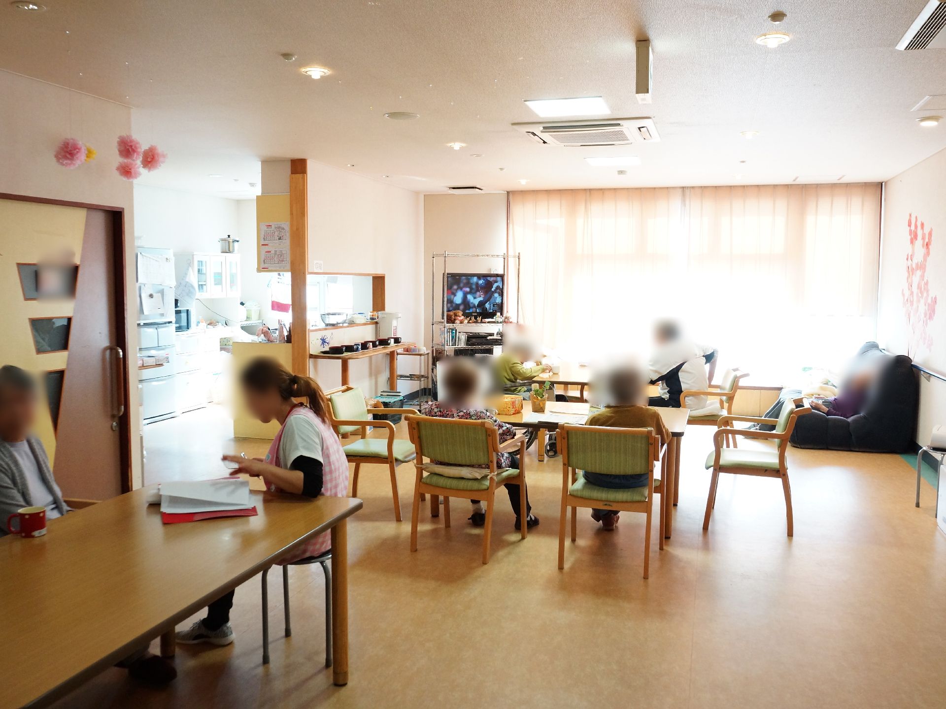 広く明るい居間で、利用者はそれぞれのんびり寛いでいる。カウンターには湯茶の用意や大型テレビも設置されている。