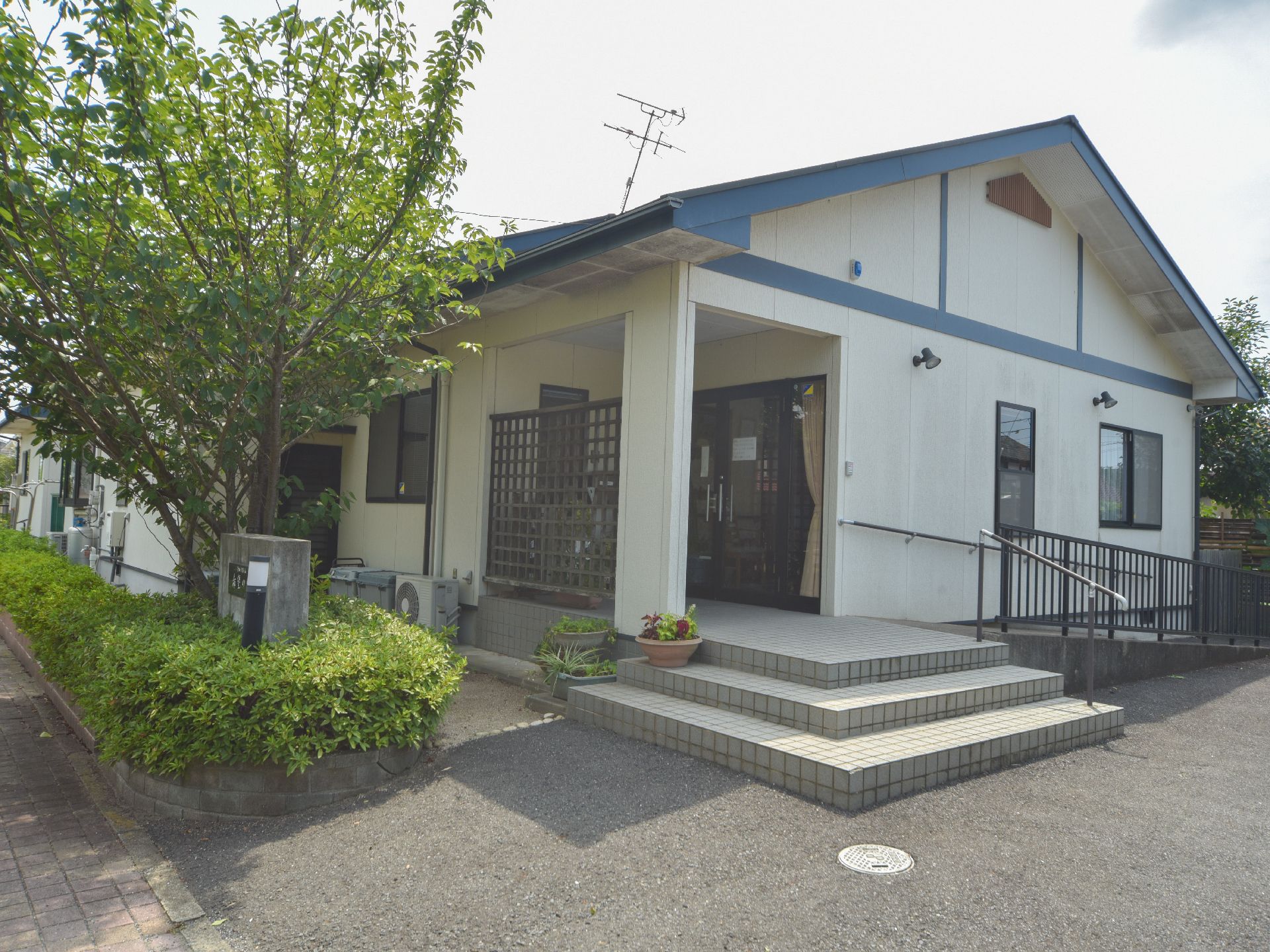 施設のエントランスまでは3段の階段と手すりのあるスロープが設置されている。施設の周辺にはたくさんの木が植えられている。
