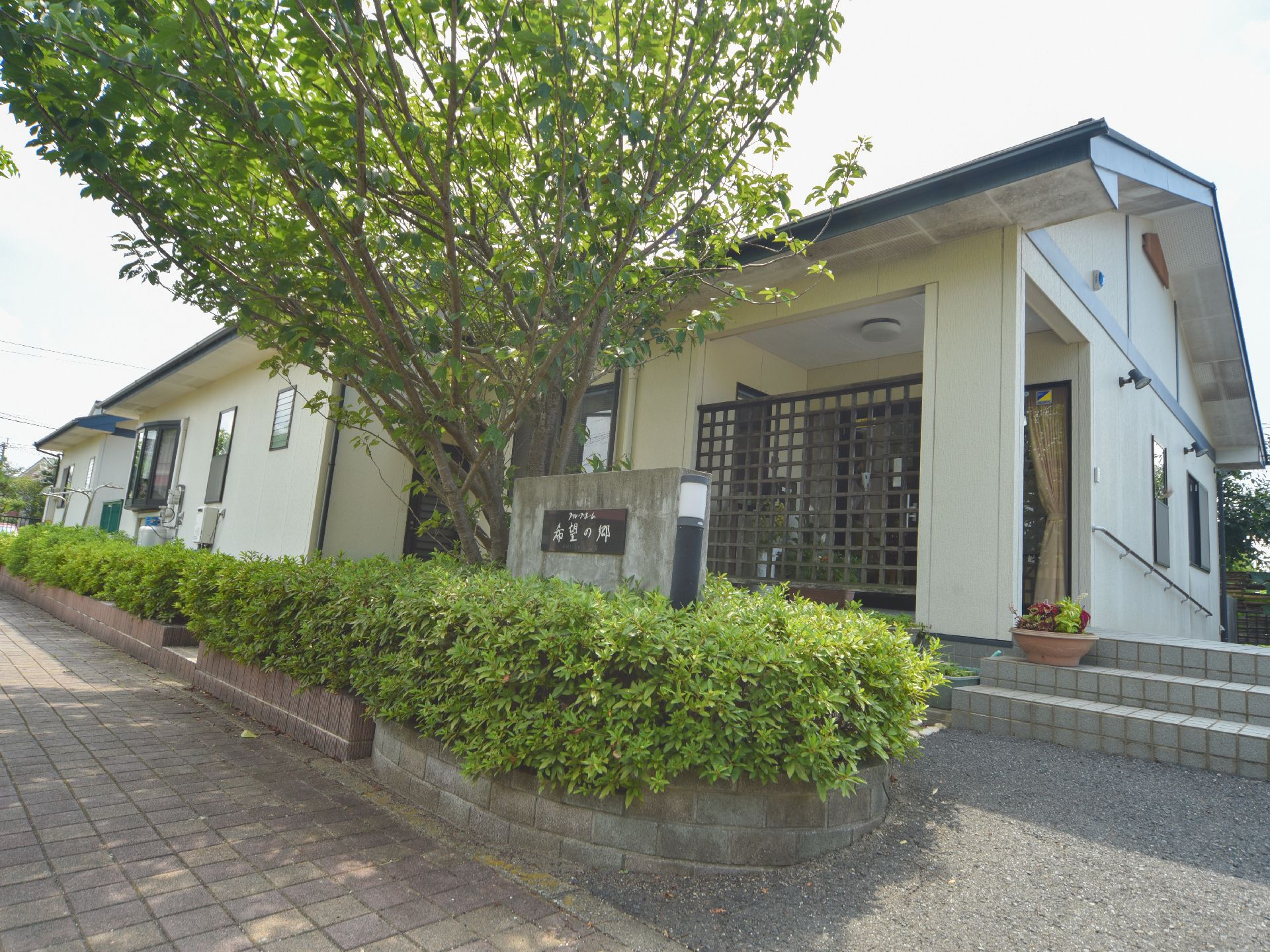 施設敷地との境の沿道に沿って背の低い木が植えられている。石に掘られた施設の名称の隣には街灯と背の高い木がある。