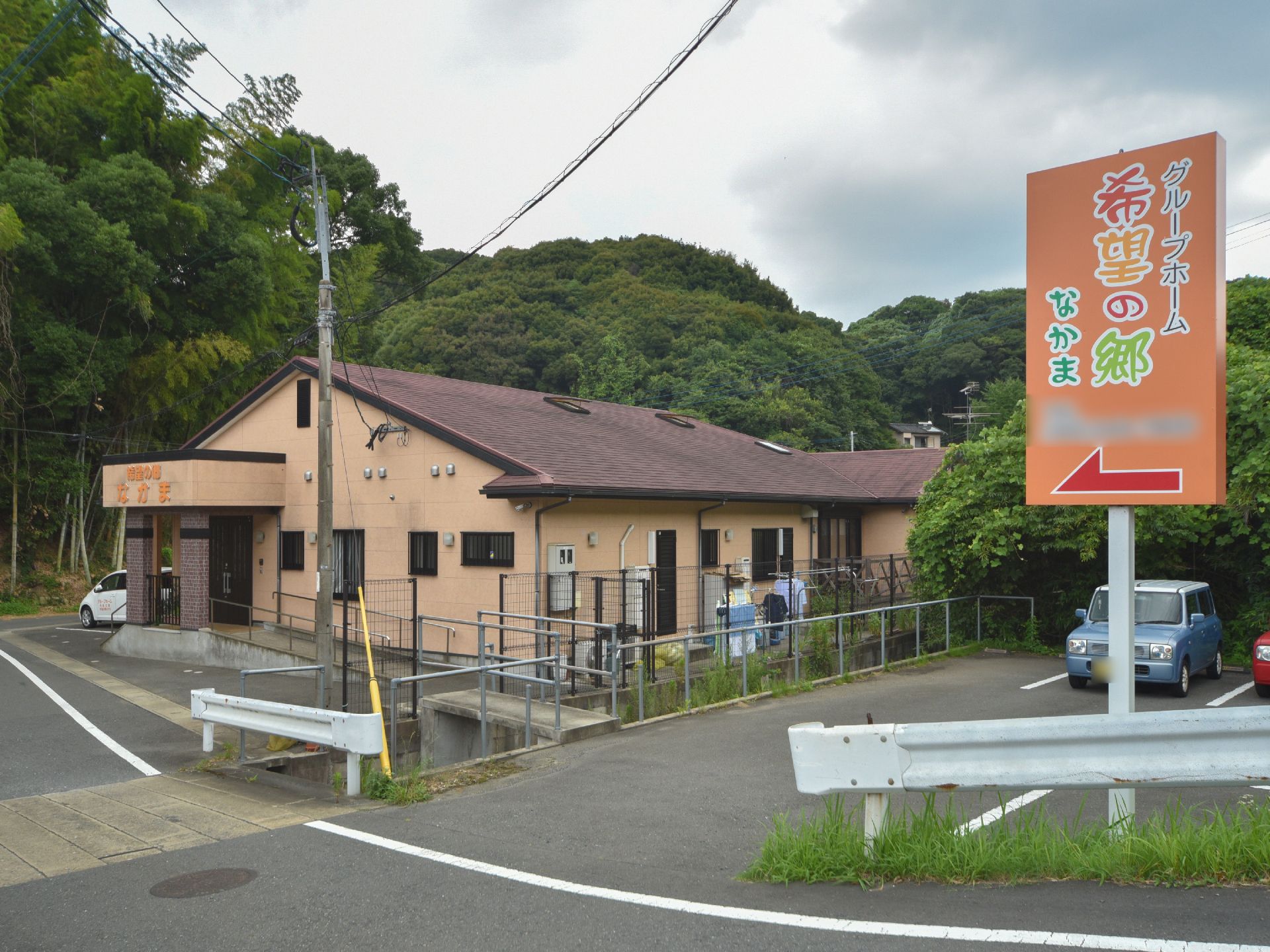 施設の敷地の奥には多くの緑があり、静かな場所に立地している。施設の少し手前に看板が設置されている。施設は1階建て。