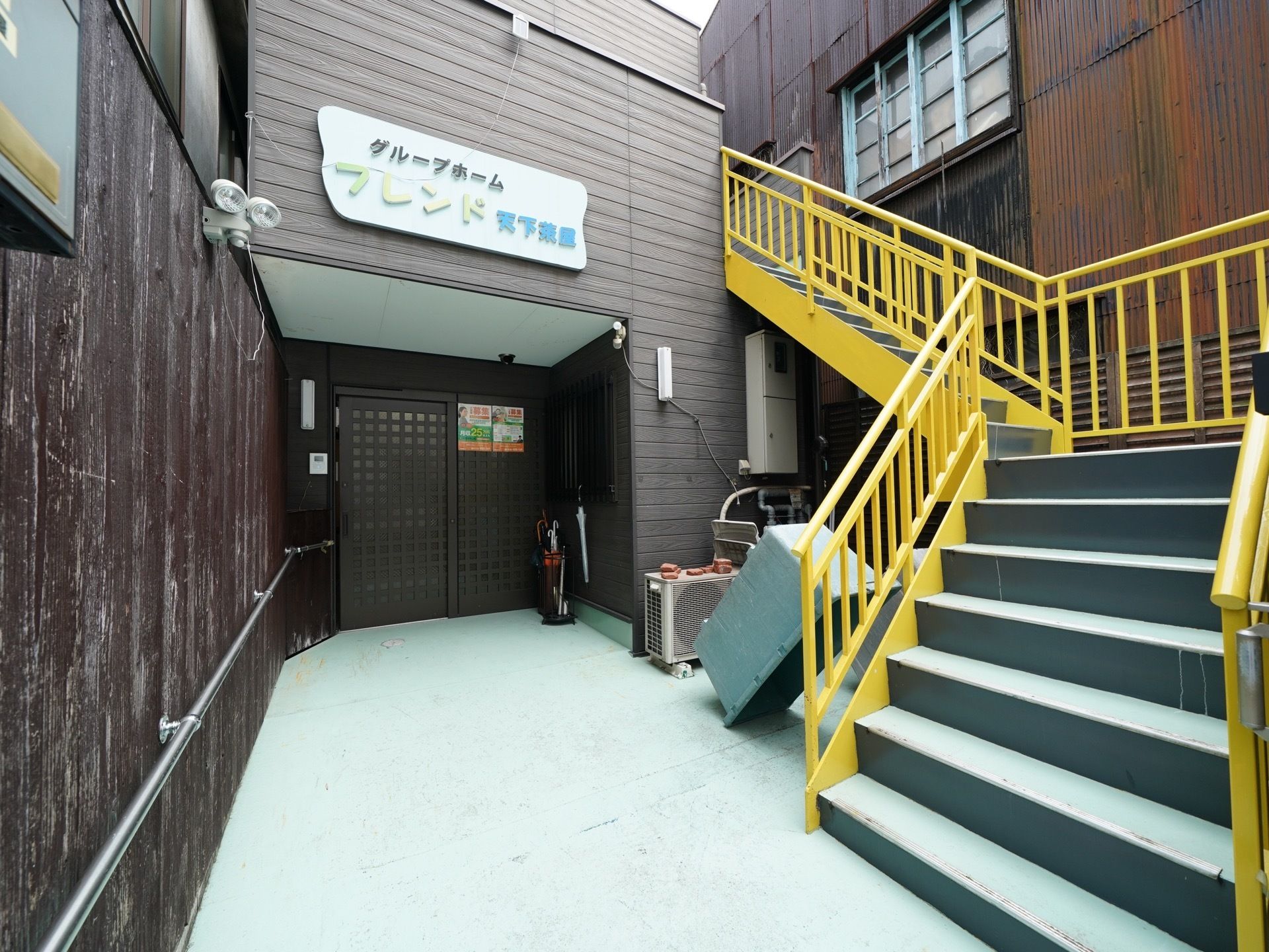濃いブラウンの外壁の建物で、出入り口までは木製の壁が設置されている。壁には手すりが取りつけられている。