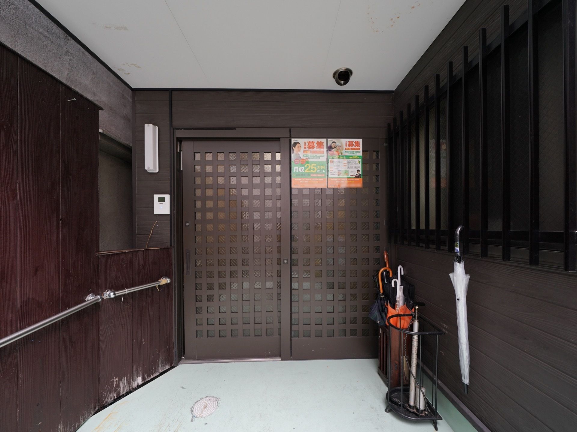 玄関には引き戸が設置されていて扉にポスターが２枚貼られている。扉の前には傘立てがあり、傘が複数入っている。