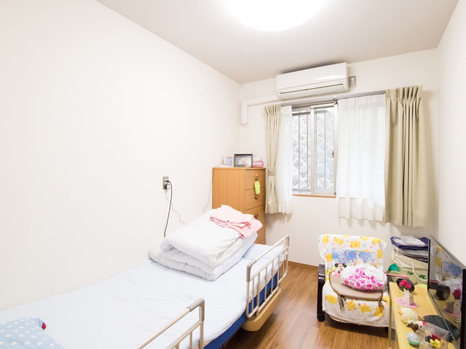 居室にはベッドとエアコン、棚、テレビなどが設置されている。ベッドには白い布団がたたんで置かれている。