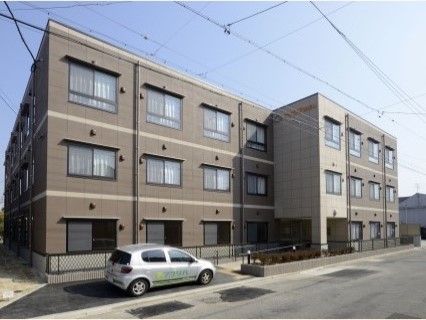 住宅街に馴染む、落ち着いた雰囲気の外観である。建物の前には植え込みとフェンスがあり、車が１台停まっている。