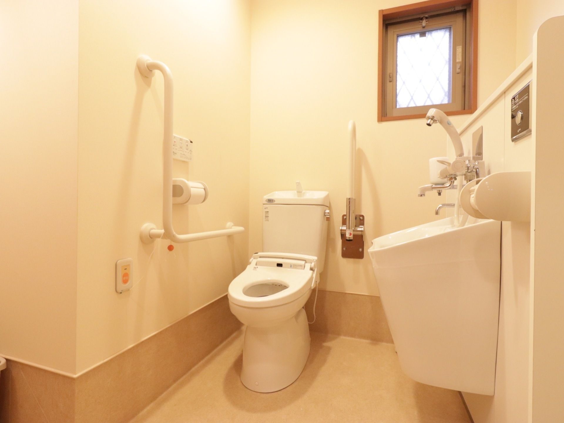 温水洗浄機能付き洋式トイレである。壁には操作パネルやＬ字型の手すり、ナースコールが取り付けられている。オストメイトに対応したトイレである。
