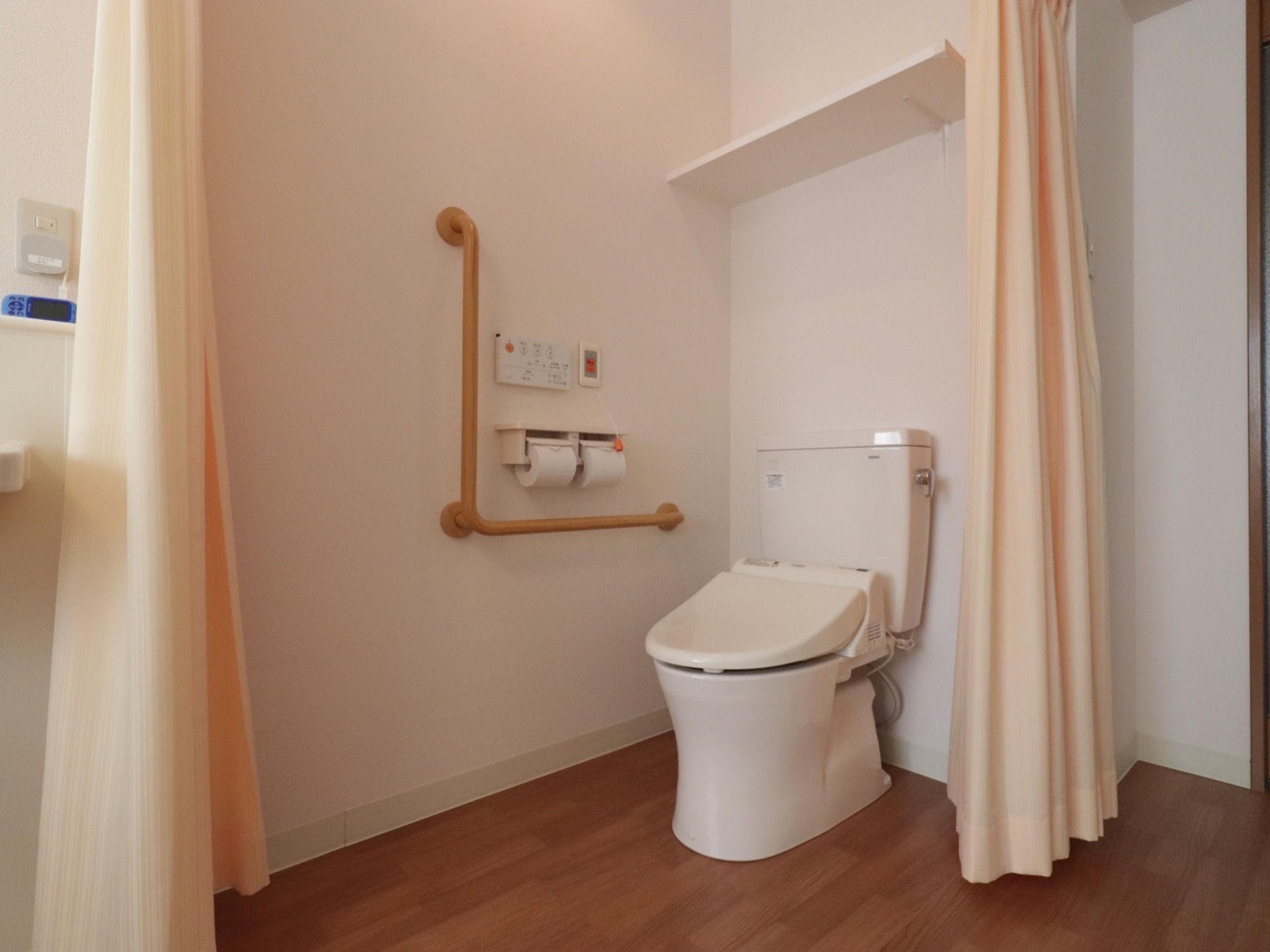 便座の近くには手すりとトイレットペーパーホルダーがある。トイレの周りには囲むようにカーテンが取り付けてある。