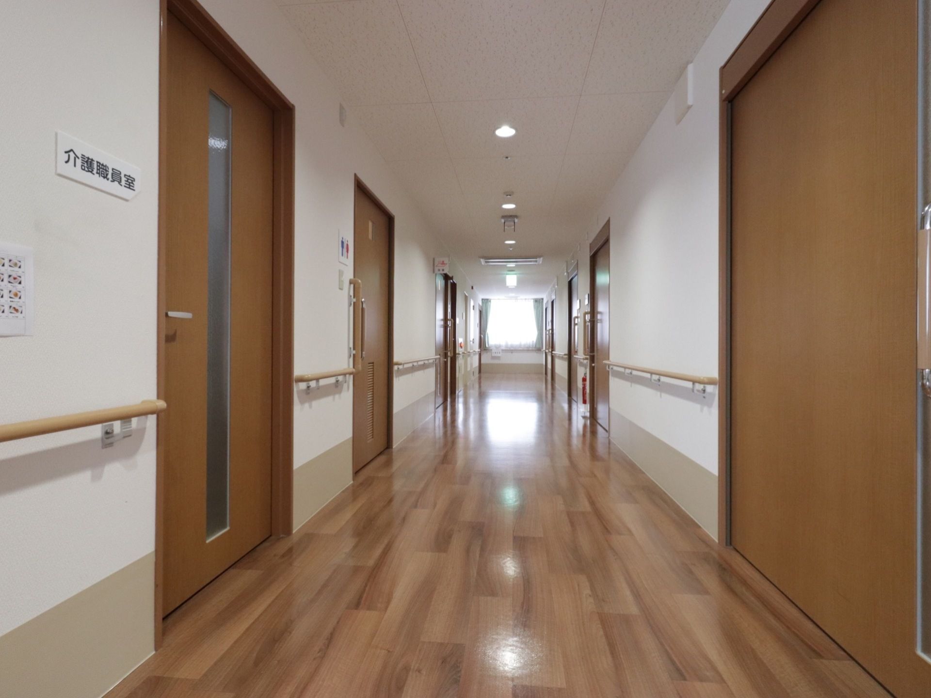 介護職員室の横にはトイレルームがある。壁と扉の近くには手すりがある。消火器が置いてあり奥には窓がある。