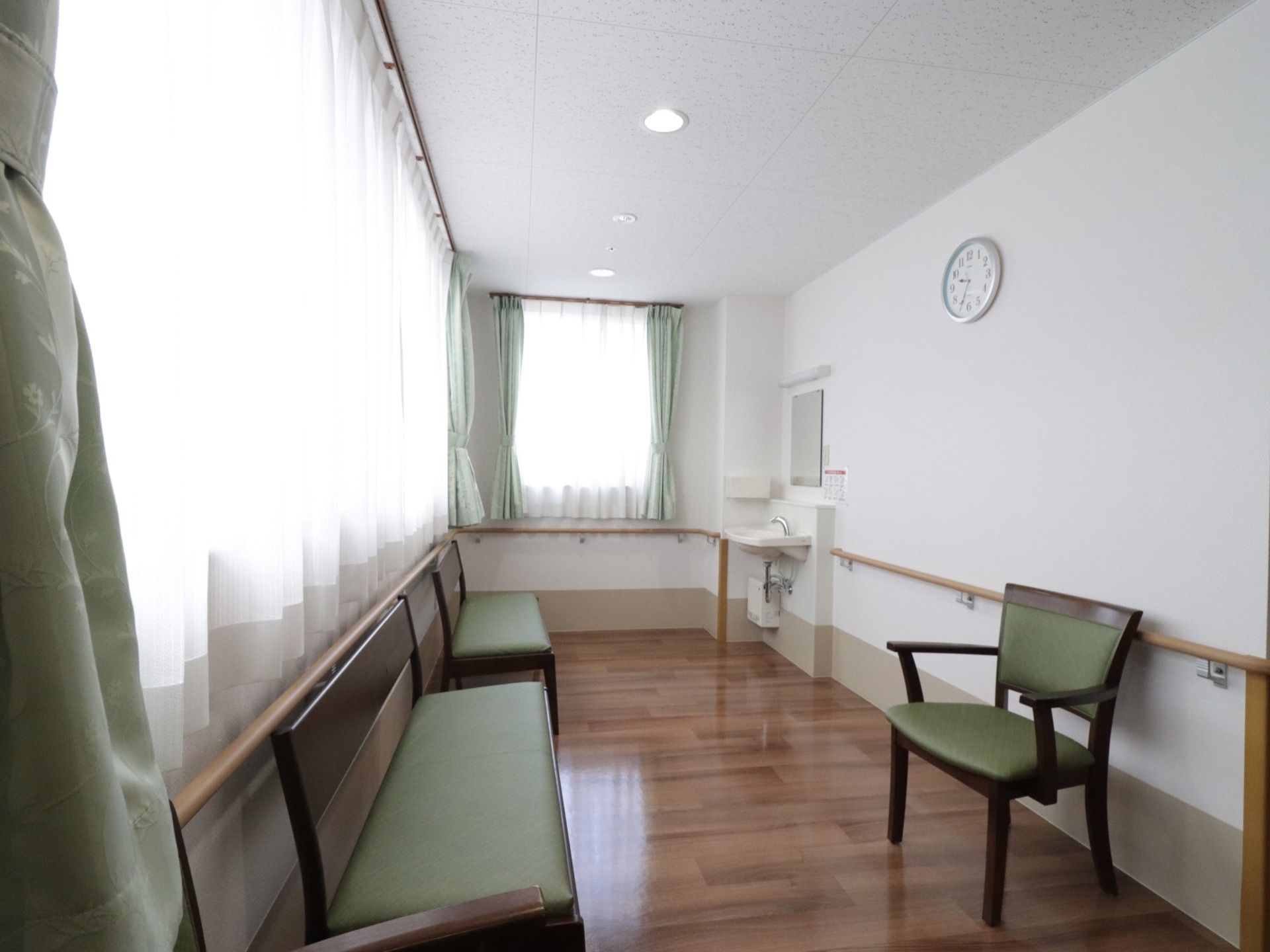 緑色の椅子が三つある。壁には時計と手すりが取り付けられている。洗面台が設置してあり、窓にはカーテンが引いてある。