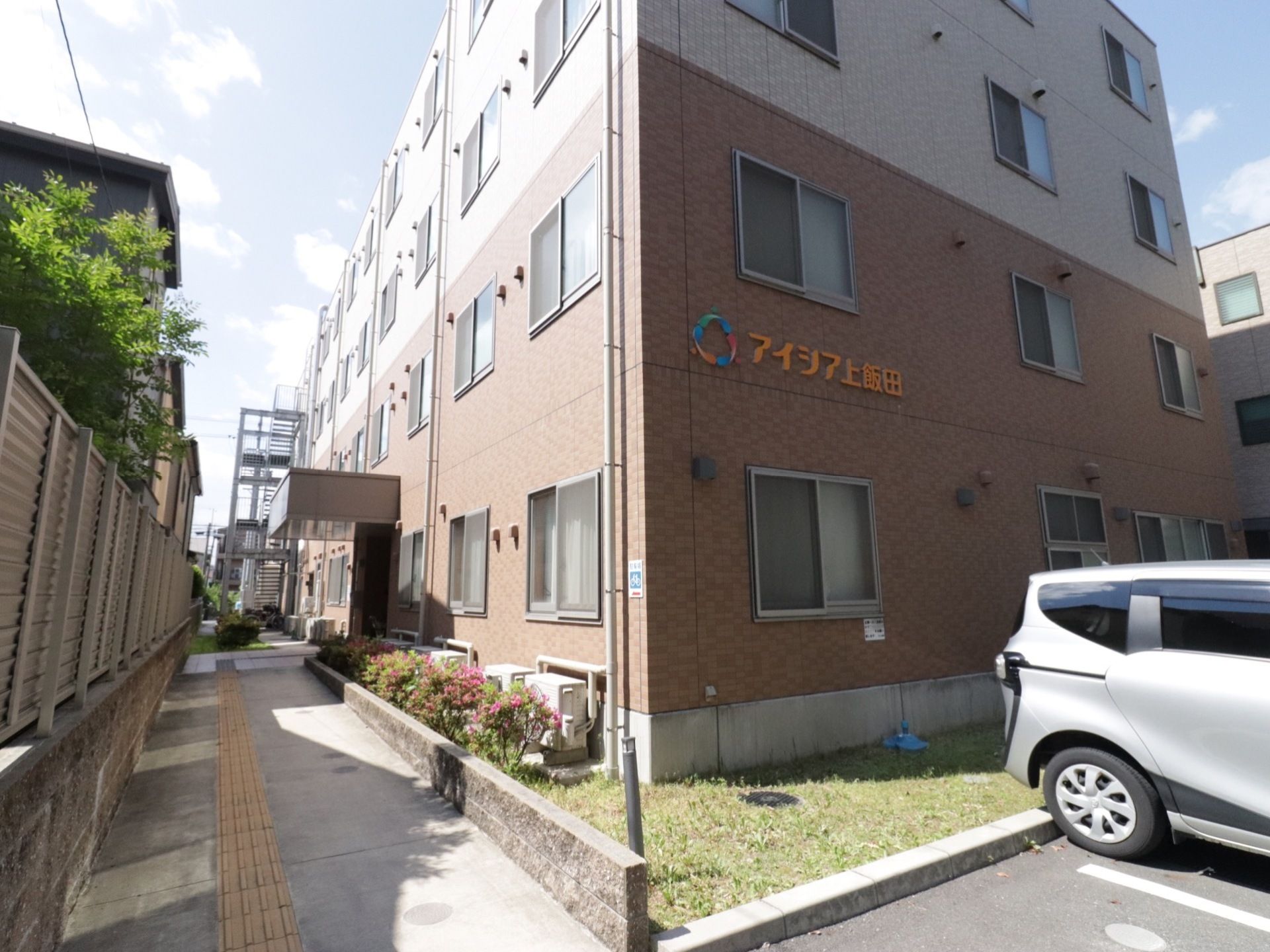 施設名が壁に表示されている。窓は均等に取り付けられている。建物の近くに駐車場があり、通路に点字ブロックが設置してある。
