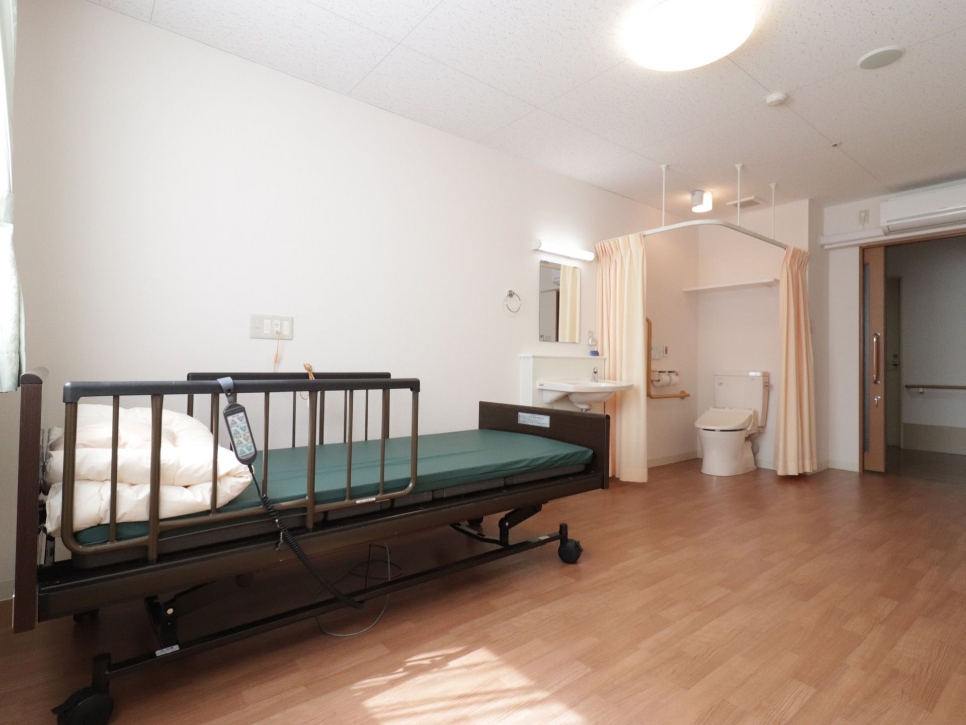介護用のベッドがある。近くには洗面台とカーテン付きのトイレがあり、天井と洗面台の上には照明が取り付けてある。