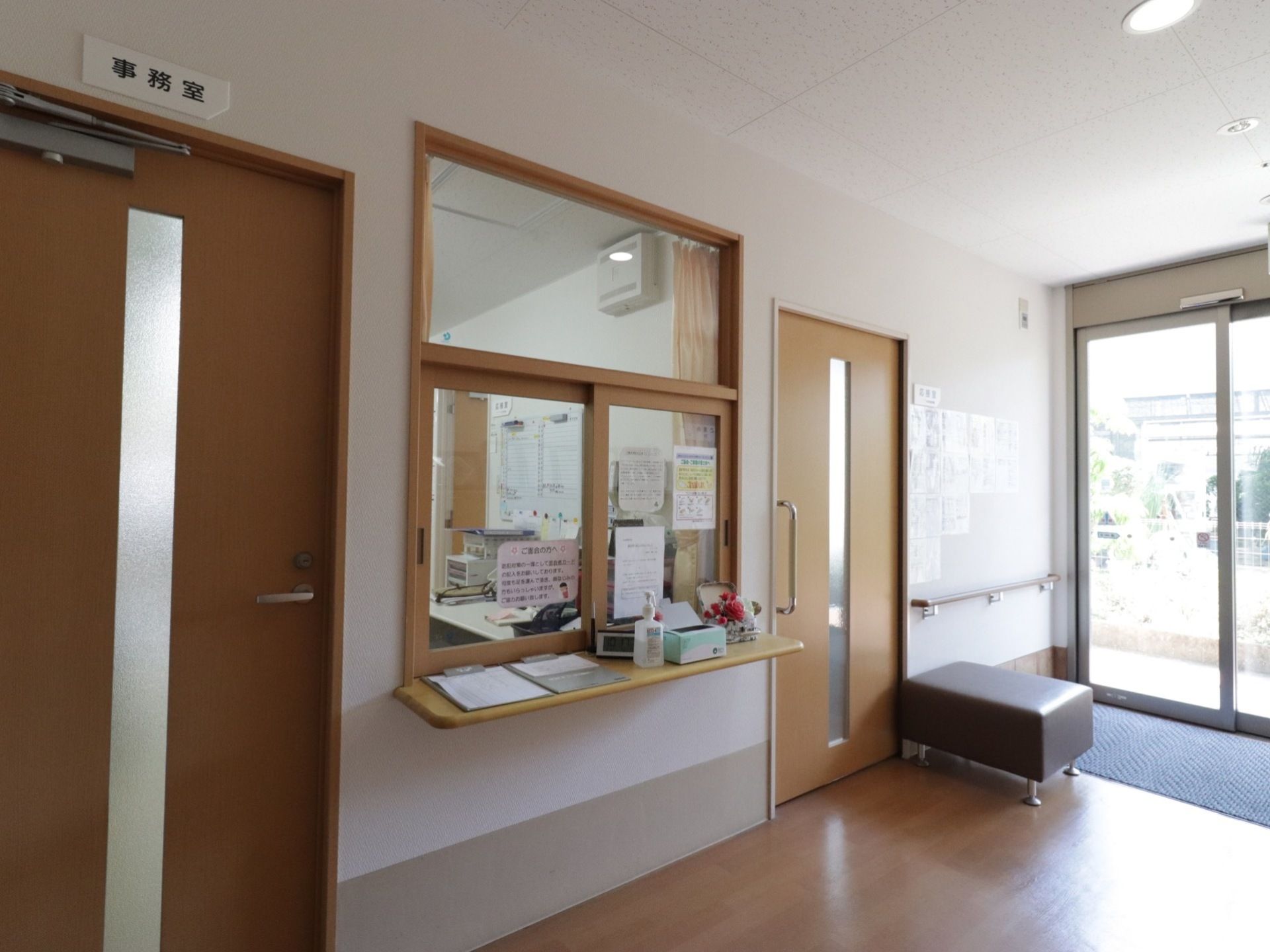ガラスサッシの自動扉が完備されている。手すりや椅子が設置され、受付カウンターの上には、来訪者様の名簿や手指消毒剤が置かれている。