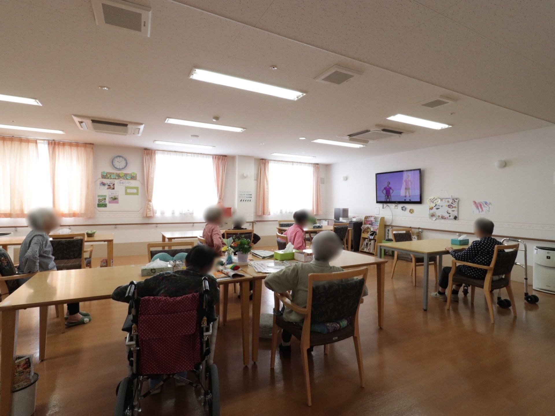 椅子やテーブル、テレビが設置されている。テーブルには花や、ティッシュが置いてある。窓があり明るく、床はフローリングになっている。