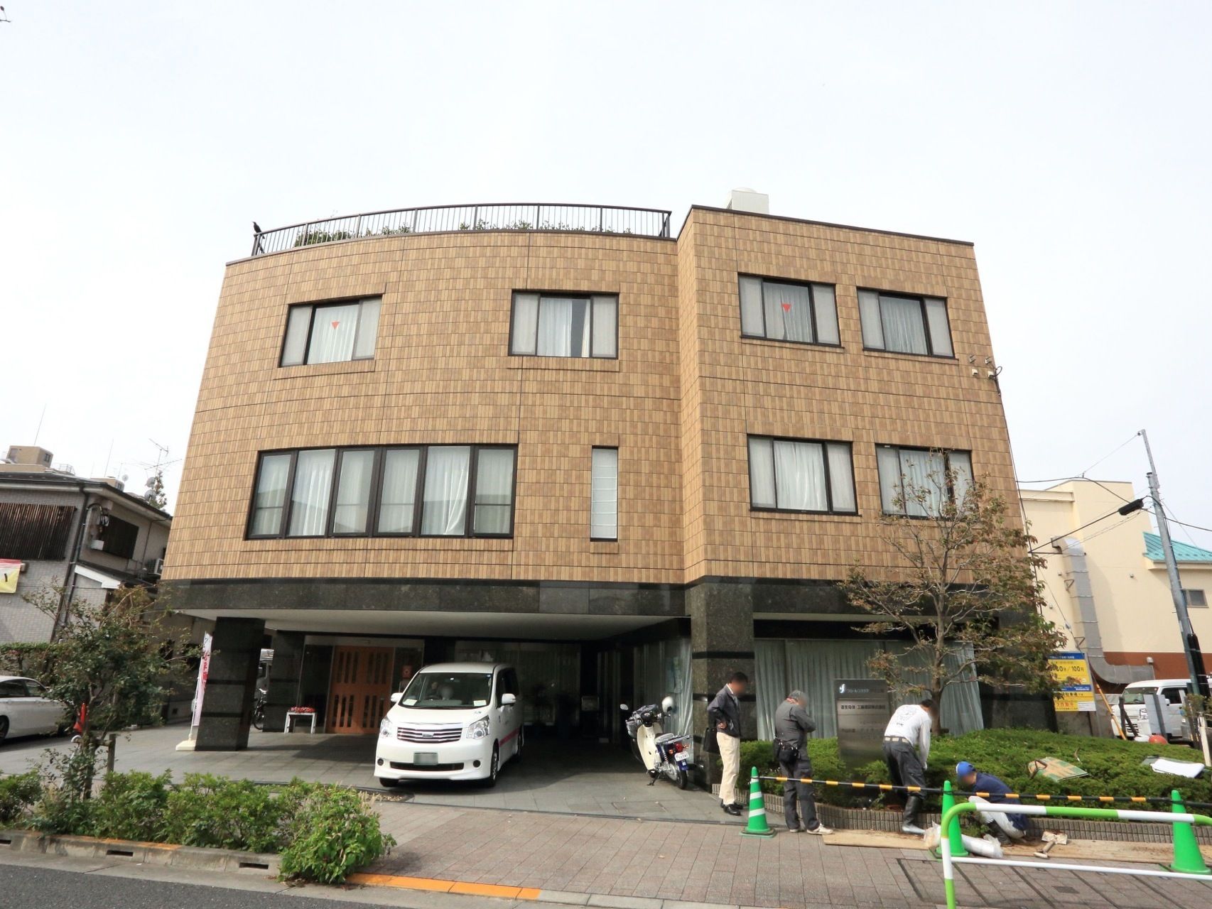 施設の一階部分には駐車スペースがあり、そこに玄関が設けられている。施設の前には背の高い木が植えられている。