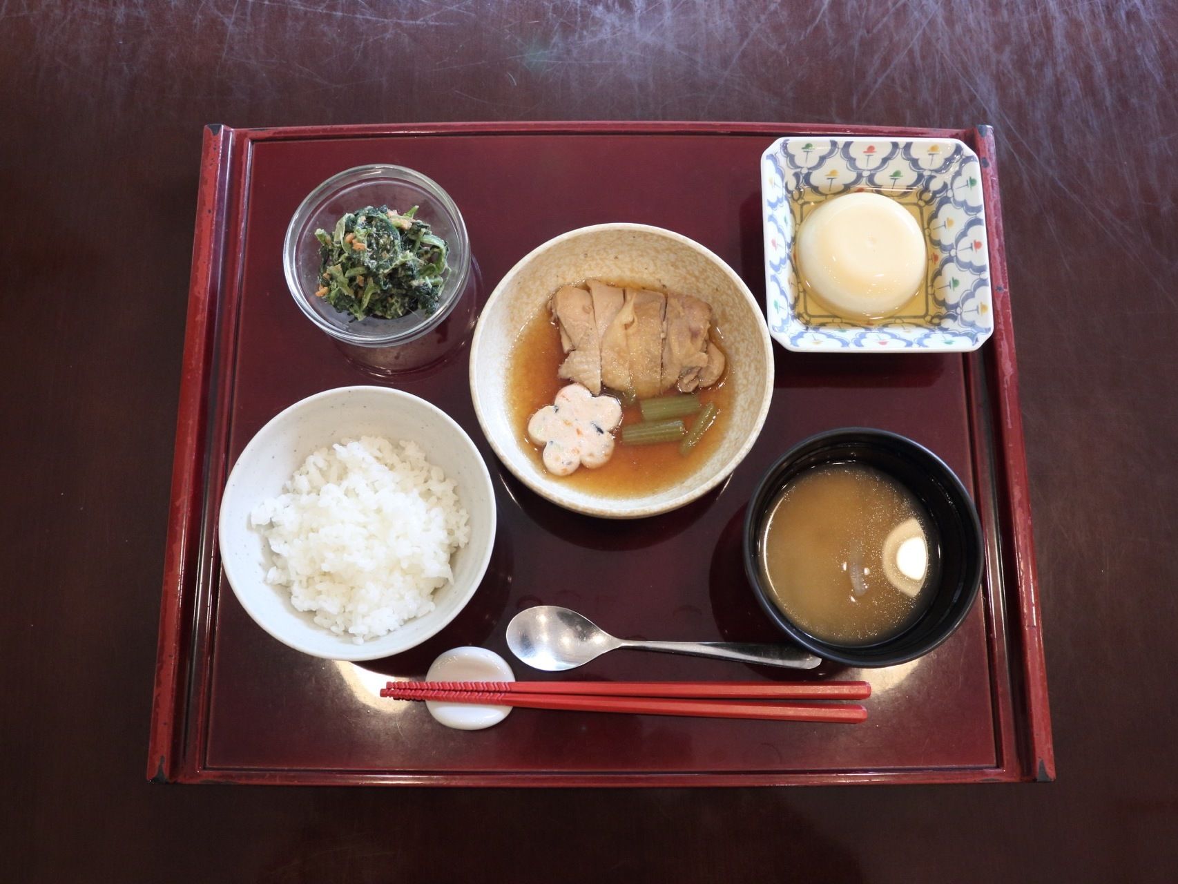 朱色のお盆の上に、ご飯、味噌汁、鳥の煮物などが並べられている。食事の道具はお箸のほかに、スプーンも用意されている。