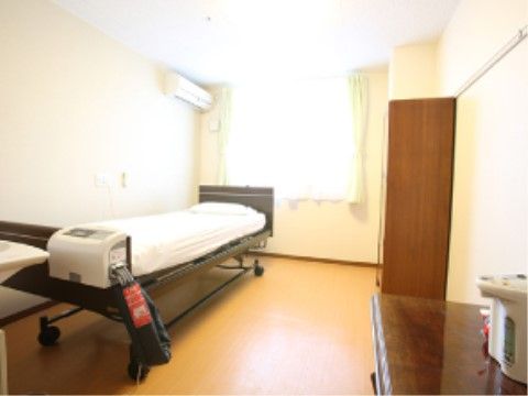 エアコンが完備された居室。キャスター付きの大きな介護ベッドが設置されており、手前の棚の上には電子ポットが置かれている。