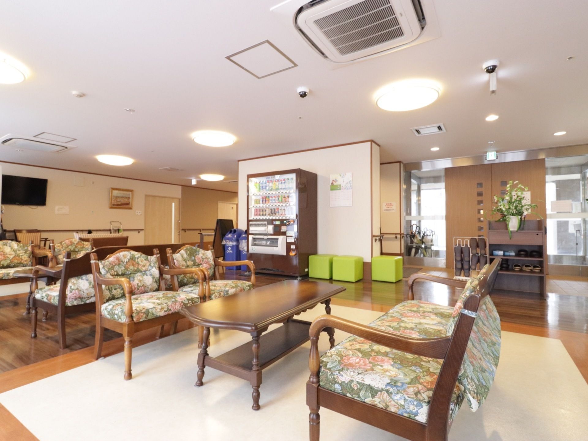 1台の自動販売機がある。近くには黄緑色の正方形の椅子やテーブル、花柄のソファーが置かれている。壁にはテレビがかけられている。