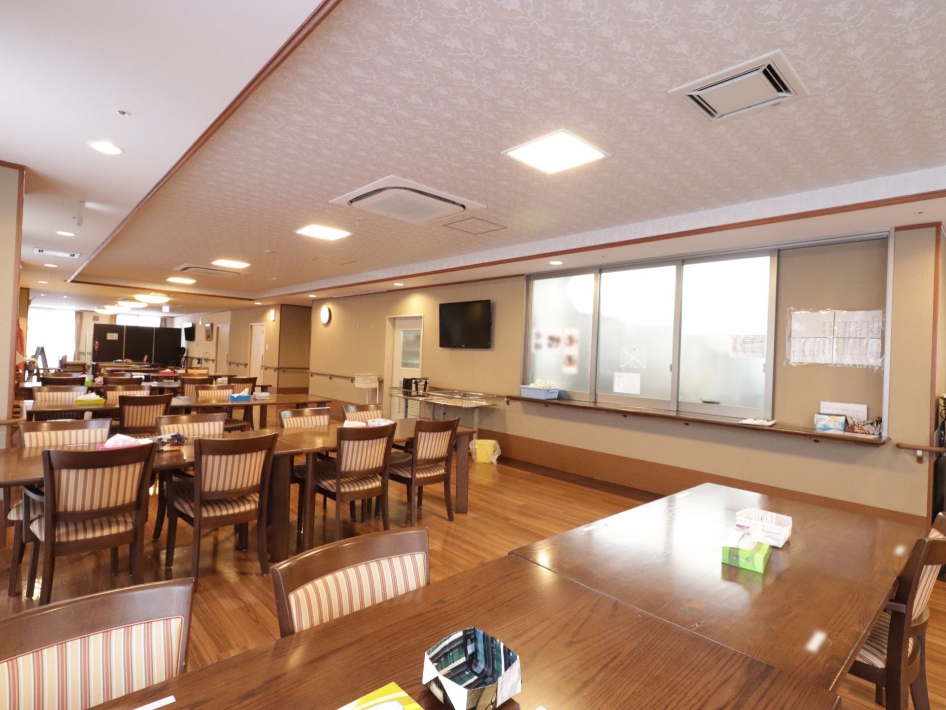 曇りガラスの窓の横の壁にはテレビがかけられている。壁には手すりが取り付けられている。テーブルや椅子が並べられている。