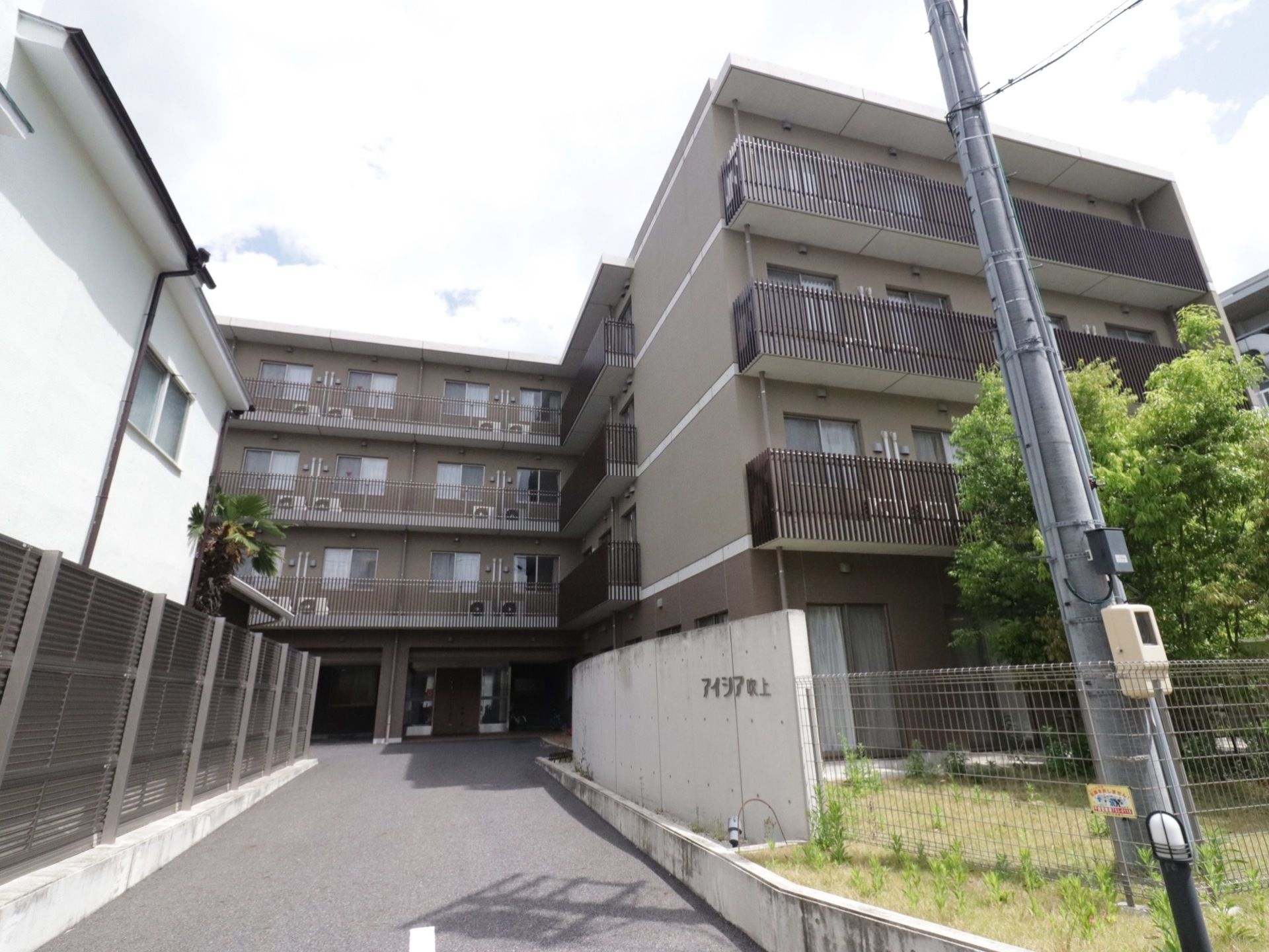 施設は4階建てである。部屋の中が見えないようにカーテンがかけられている。電柱近くにはフェンスがあり、その隣には施設名が書かれた塀がある。