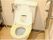 トイレは洋式便座となっている。便座の横にトイレットペーパーホルダーと手すりが設置されている。木目調の床が明るい雰囲気を漂わせている。
