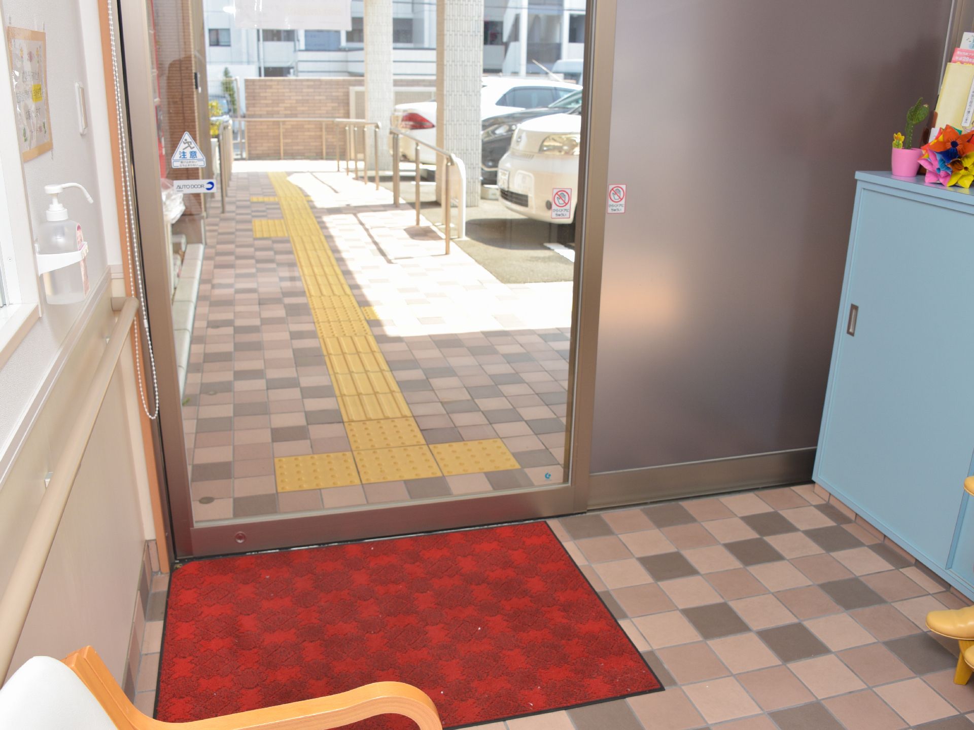自動ドアの玄関である。赤いマットが足元に敷かれている。壁際にはパステルブルーの靴入れが設置されている。