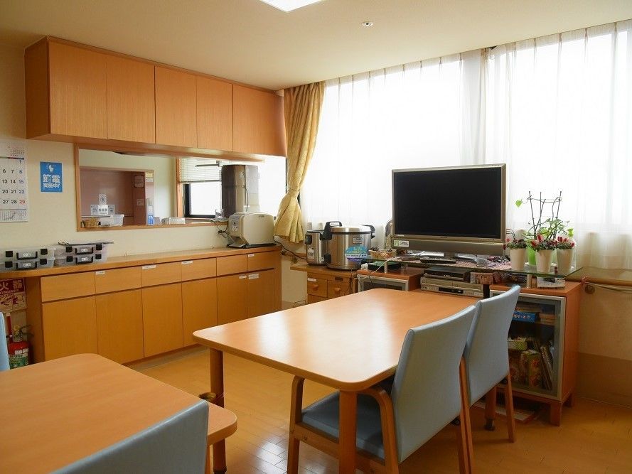 リビングには茶色いテーブルと水色の椅子が並べられている。横の棚にはテレビや観葉植物が置かれている。隣にも棚があり、そこには炊飯器がある。