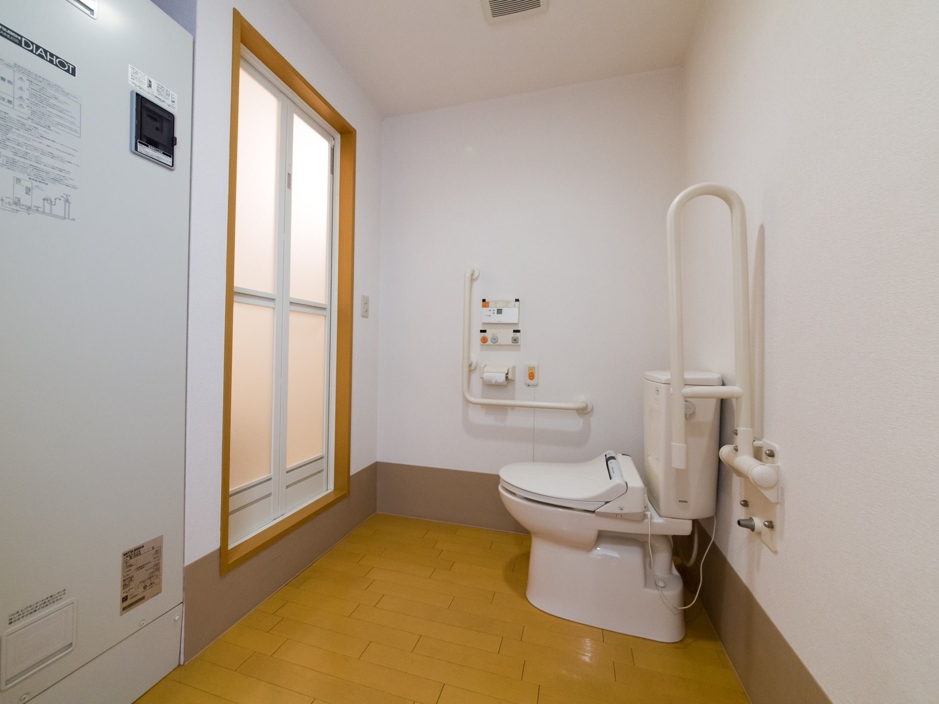 お風呂の前がトイレになっている。可動式の手すりが付いて、支えながら立ち座りをして用を足すことができる。