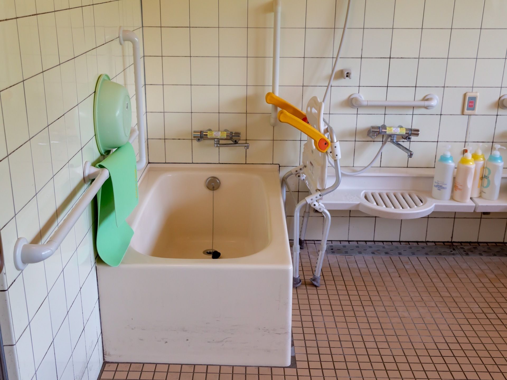 壁に手すりや白いユニットバスが完備された清掃後の浴室である。バスチェアーやすべり止めマットが水切りされている。