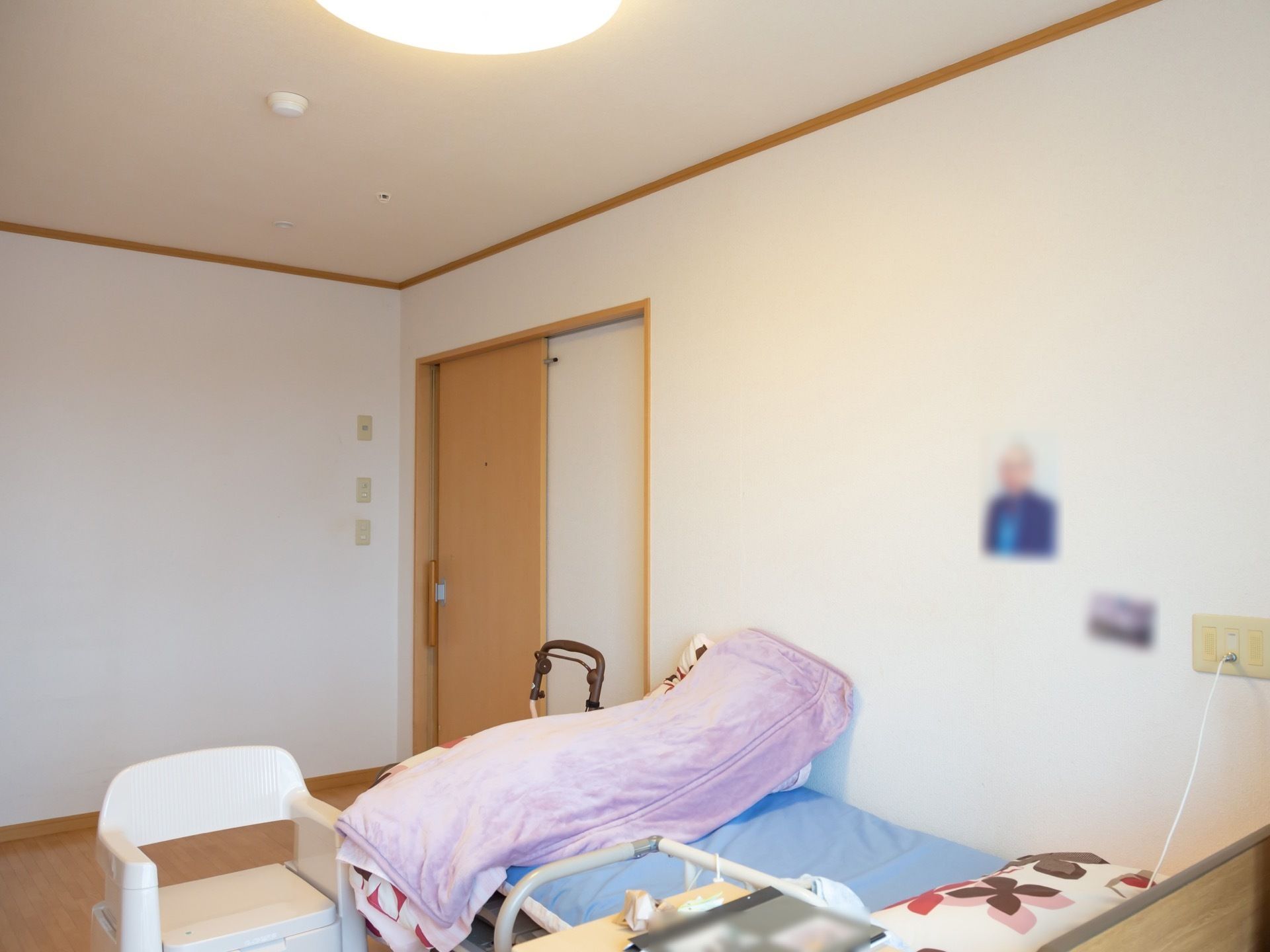 居室は明るい木目調のスライド式扉が完備されている。介護用ベッドや呼び出しブザーが設置されており、歩行器が置かれている。