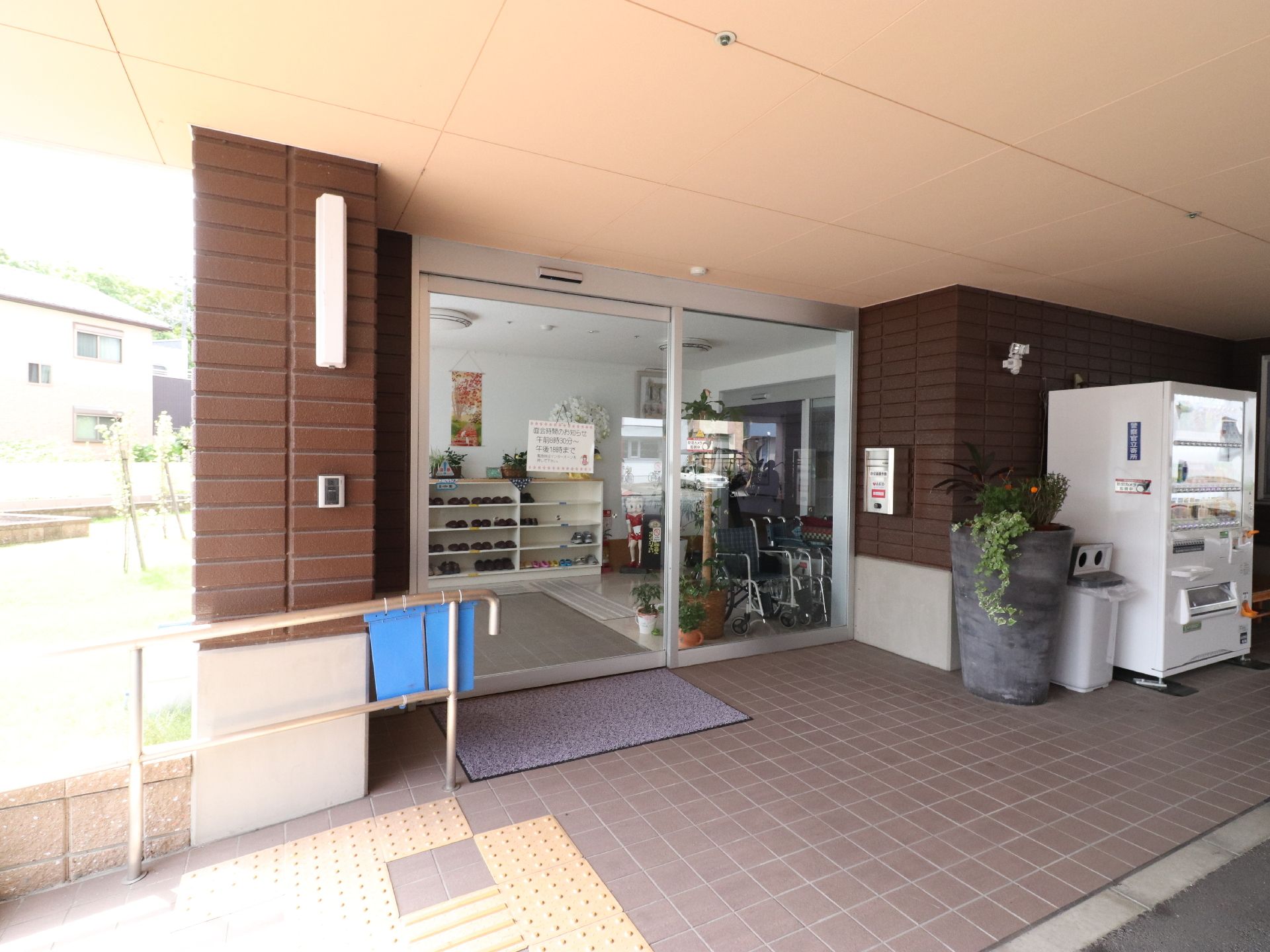エントランスにある自動ドアの周りは、緩やかなスロープがある。右手には自動販売機とゴミ箱があり、観葉植物も置かれている。