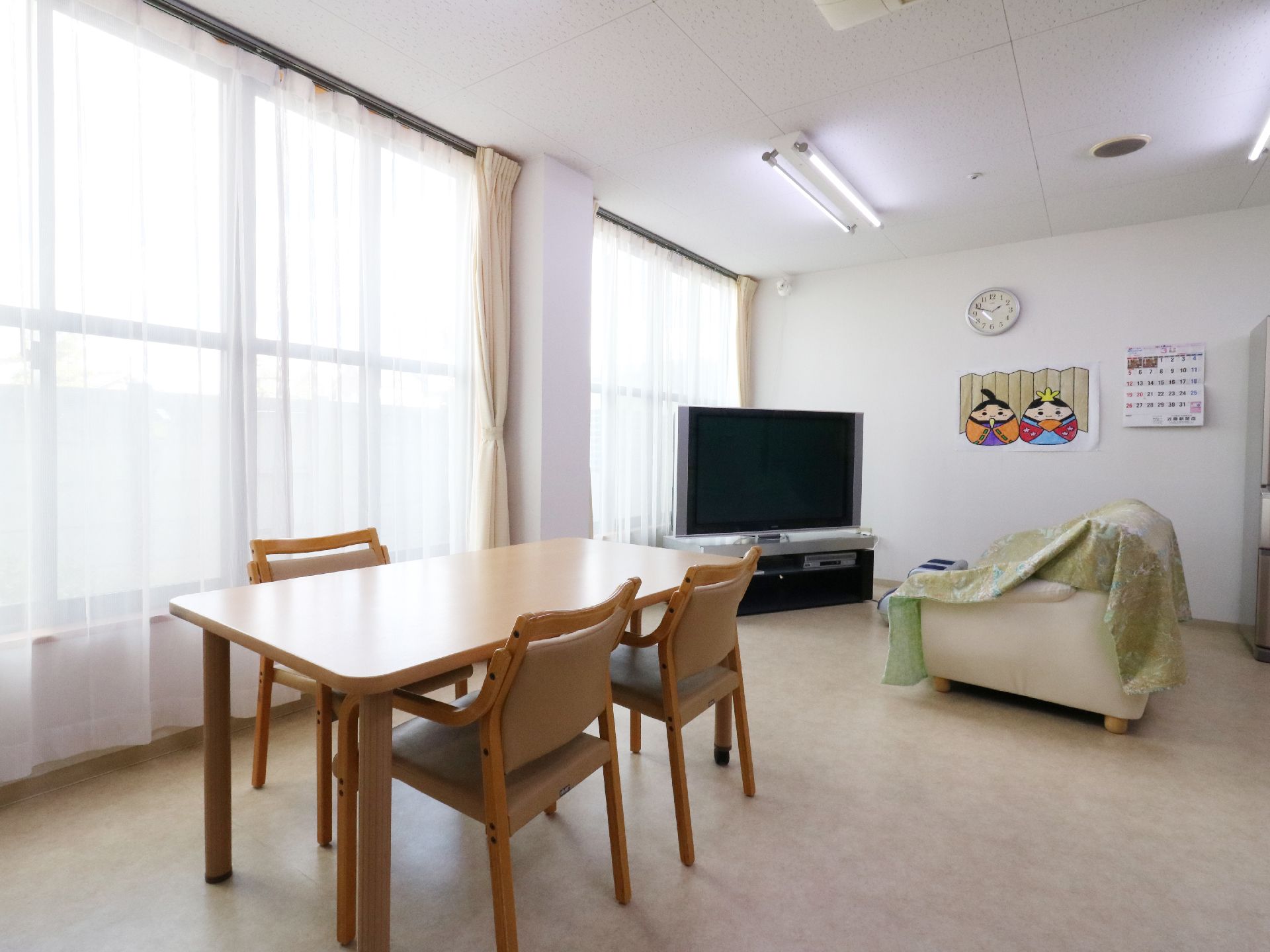 ダブルカーテンが掛けられた大きな窓。部屋の角にはテレビが設置され、その前にはソファーがあります。テーブルと椅子の場所もあります。