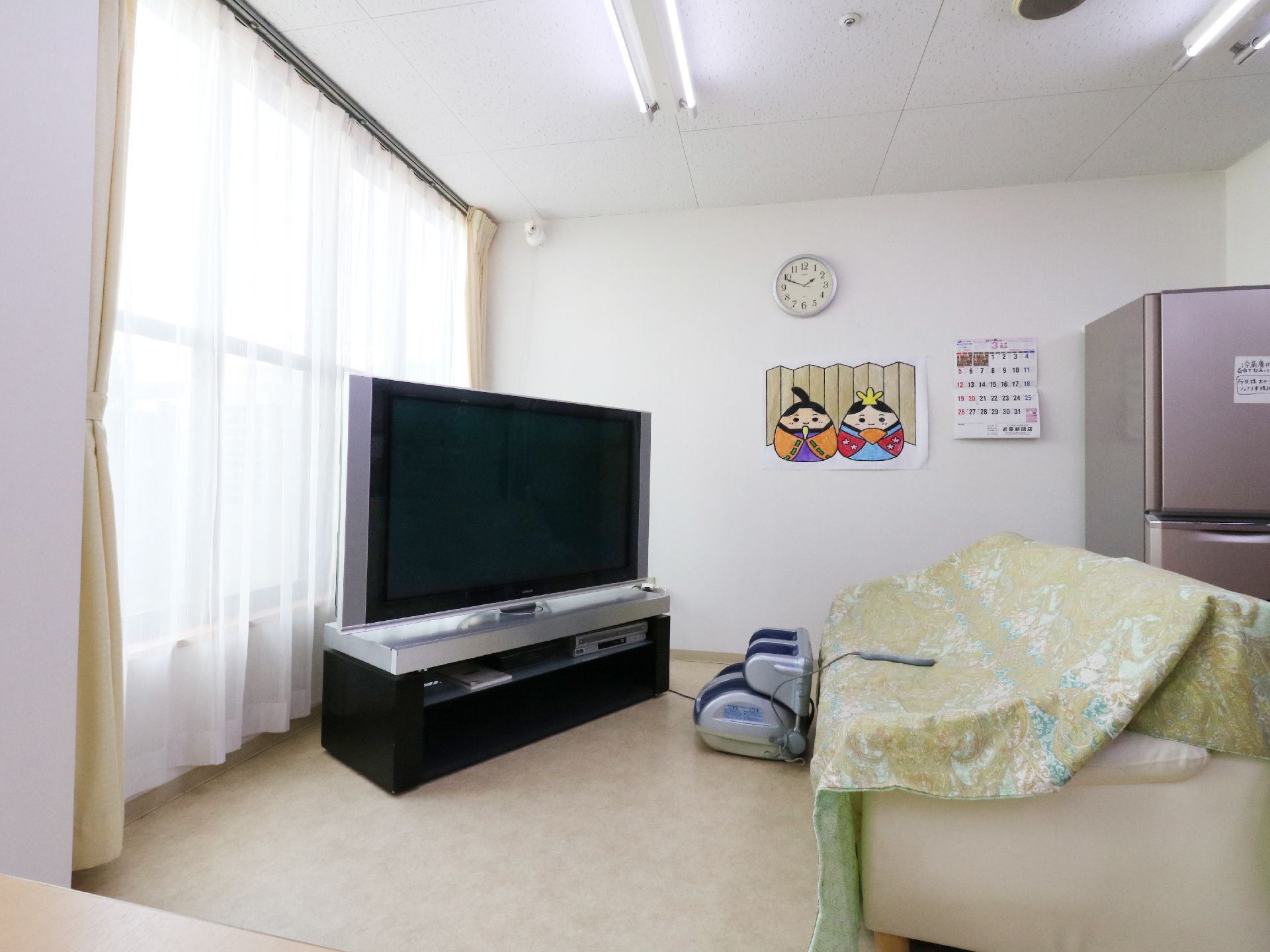 大きな窓を背にしてテレビが設置されています。その前には大きな布を掛けたソファーがあります。マッサージ器が置かれています。
