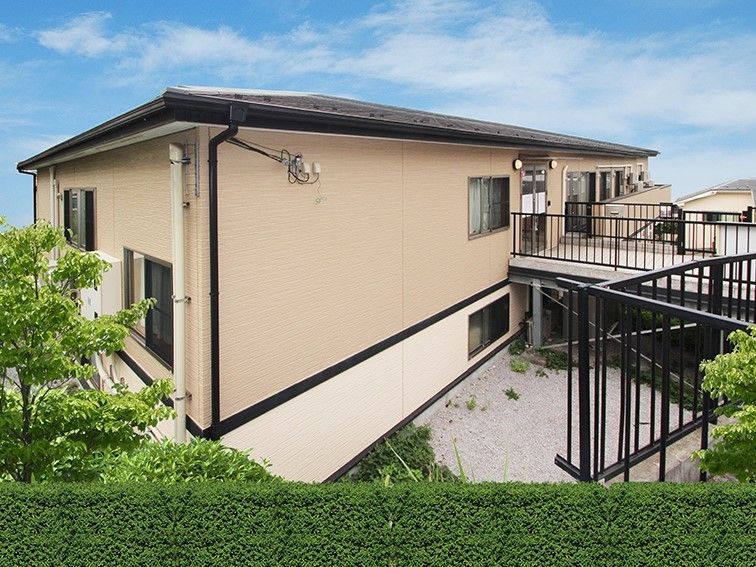 大きな屋根が付いたベージュの建物の周りには緑が多く、玄関から出た道の両サイドに手すりが付いている。