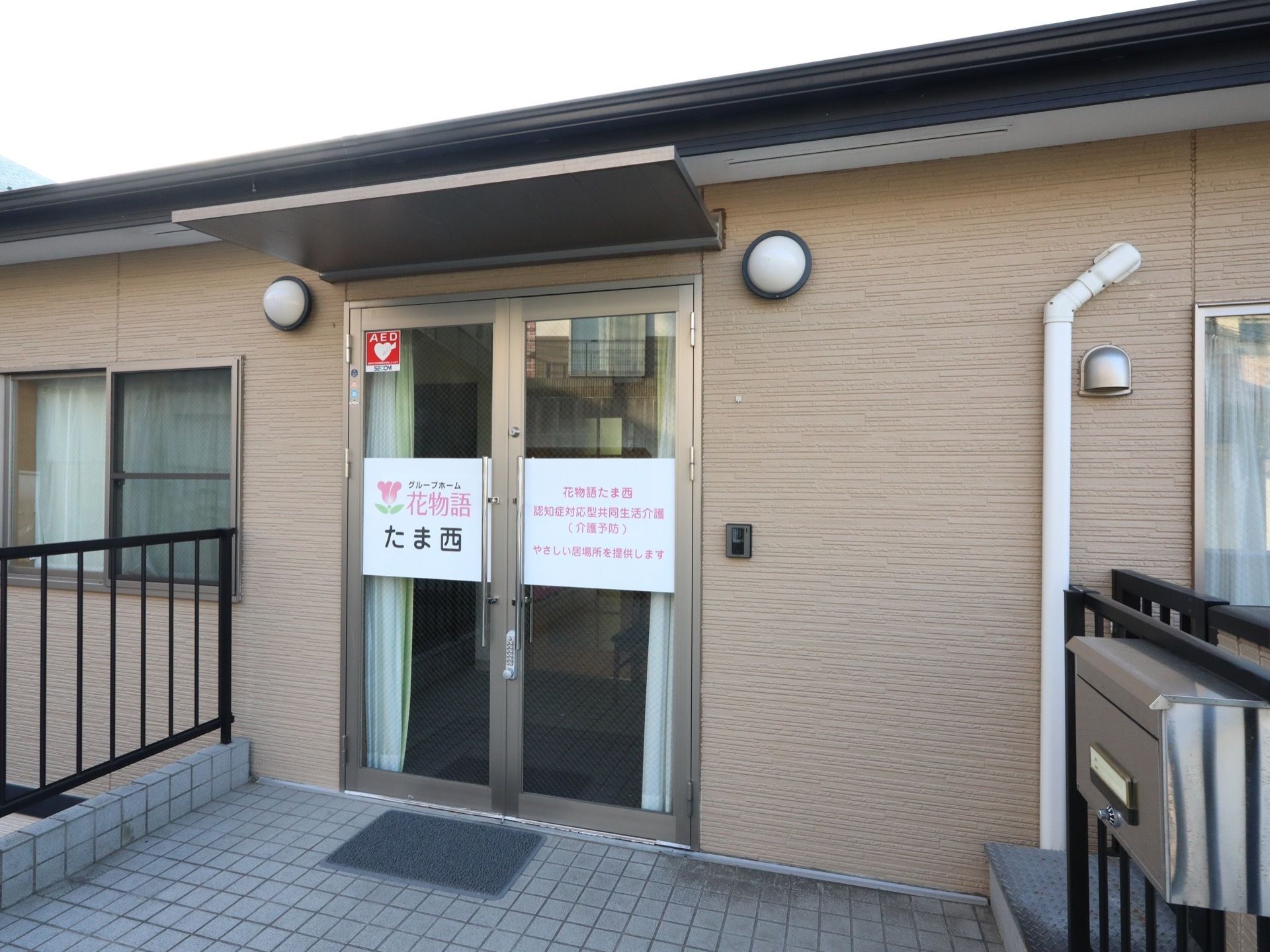 施設の玄関には、ガラス張りの２枚扉が備わっていて、施設名が書かれている。すべり止め用のマットや郵便ポストがある。