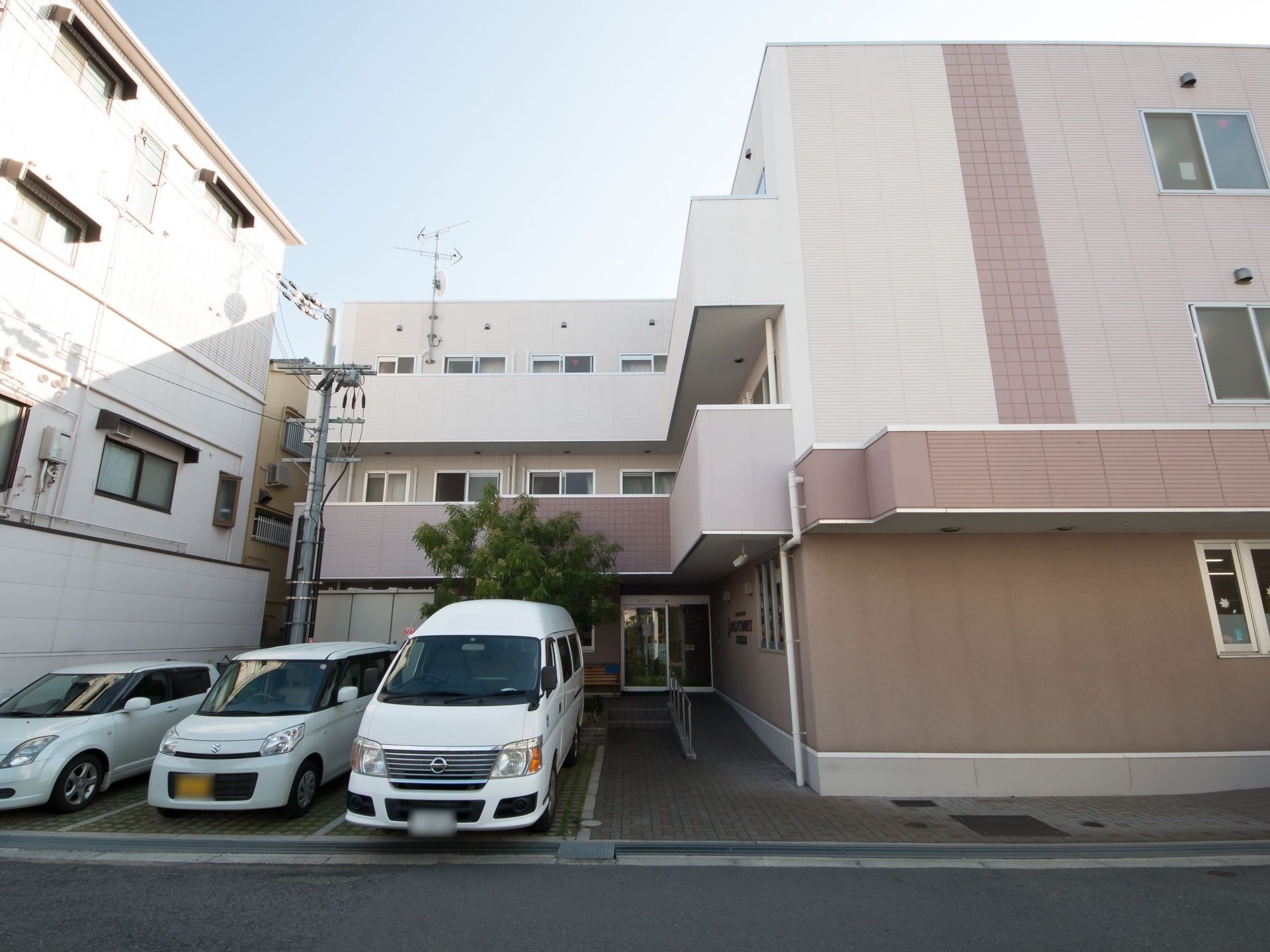 ぬくもりのある色合いの施設建物である。施設手前には駐車スペースがある。玄関前は、階段とスロープを分ける手すりが設置されている。