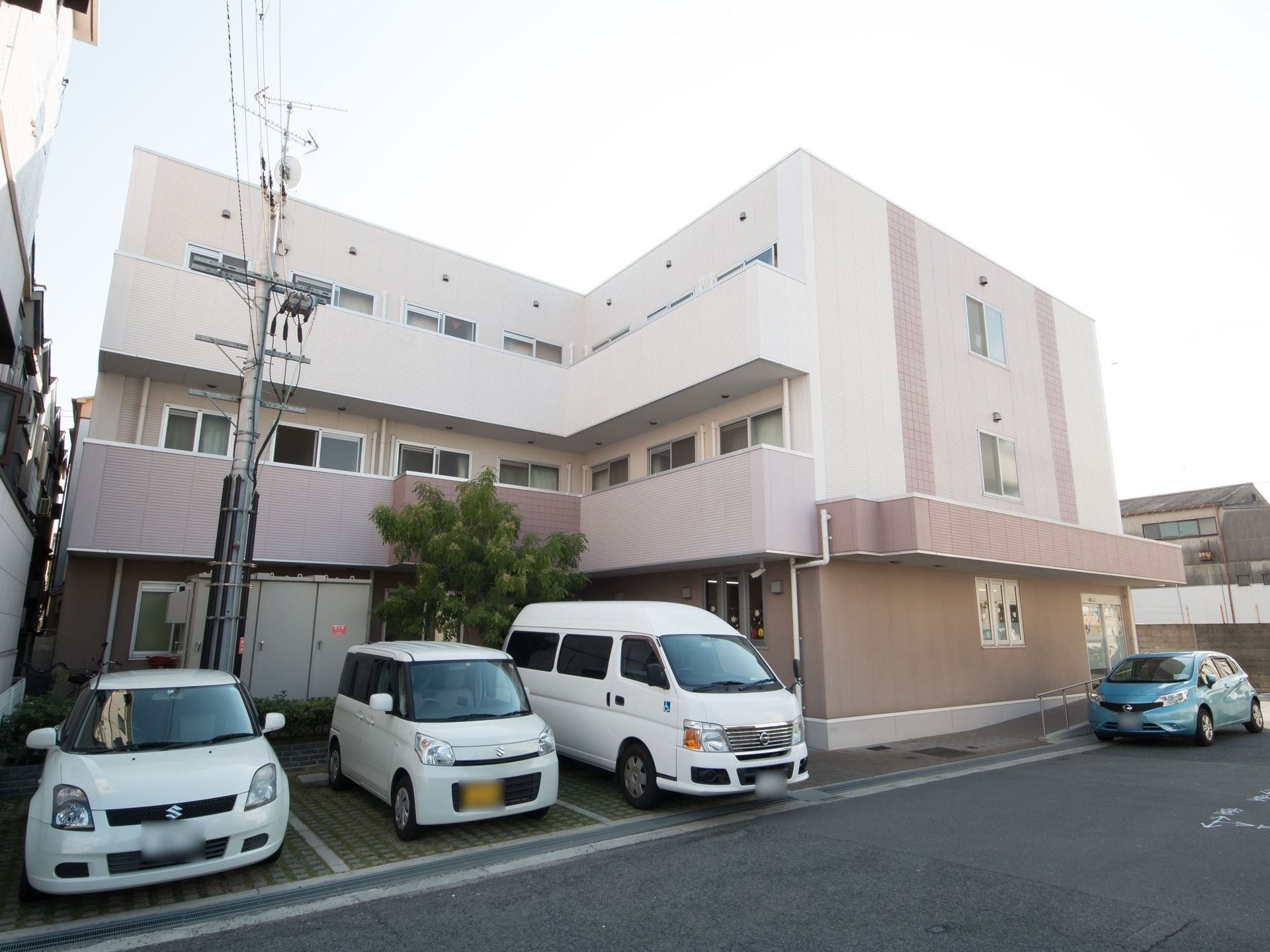 ３階建てのＬ字型の建物で、白と茶色に塗られている。建物の前に施設で所有するワゴンや、軽乗用車が停められている。