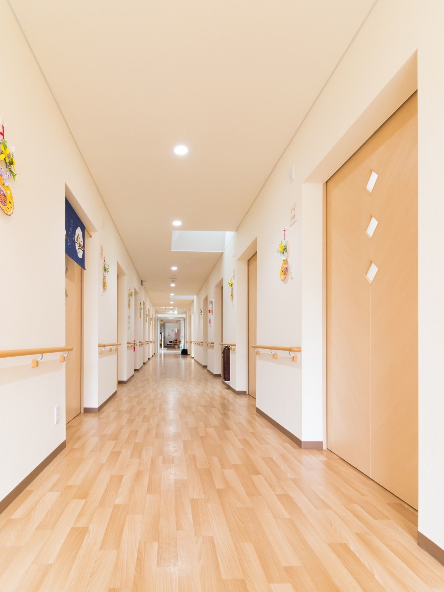 通路の両方の壁には居室扉と同じ色合いの手すりが続いている。壁には手作りの飾りが飾られている。天井にダウンライトがある。