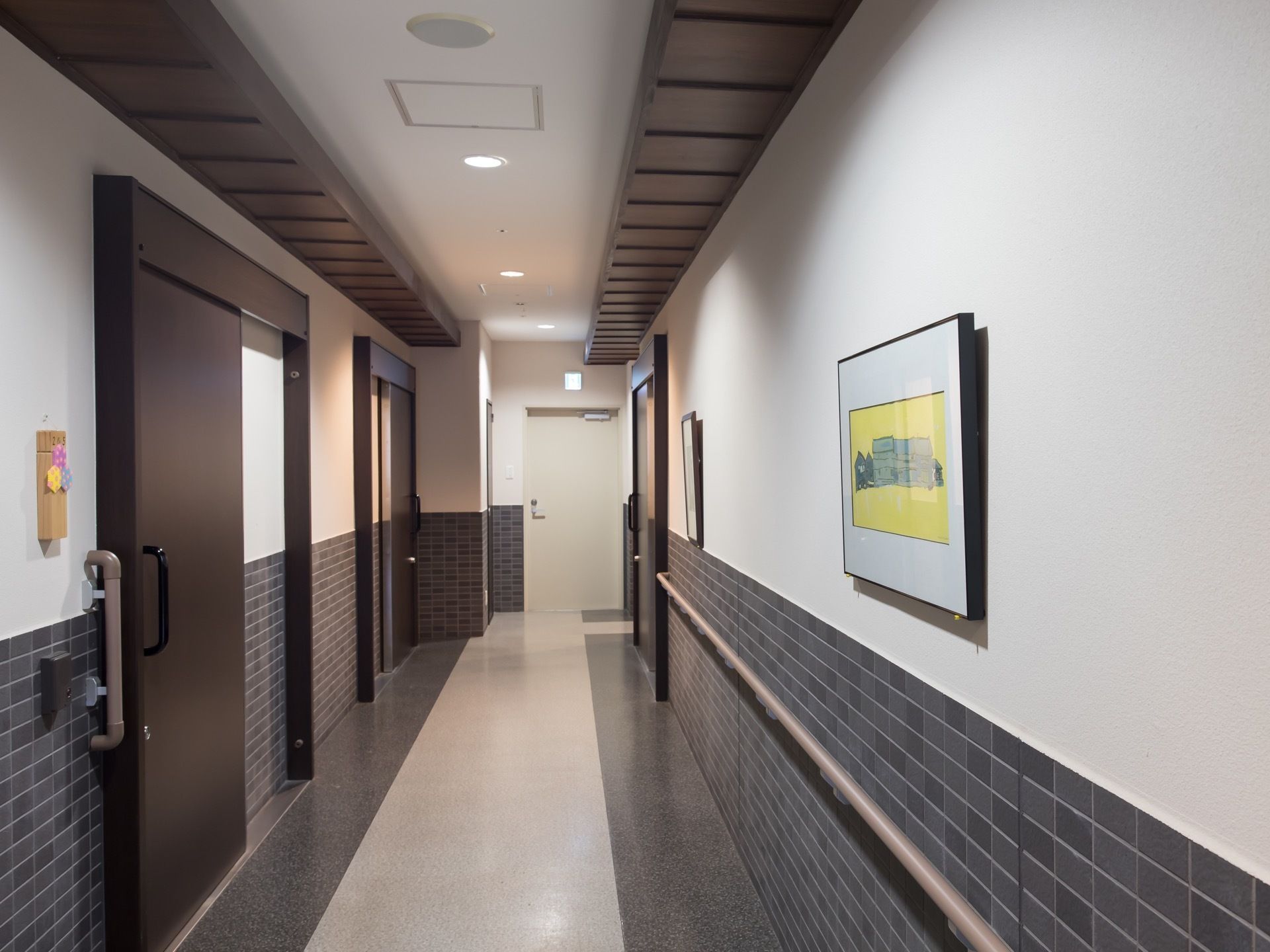 ダークブラウンの部屋の扉が並んだ通路がある。扉の正面には絵画が２枚飾られている。手すりがある。