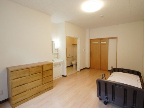 居室扉は引き戸式で、入るとすぐ横にトイレがある。温水洗浄便座の上に棚が設置されており、横には手すりが付いている。壁際にベッドが置かれている。