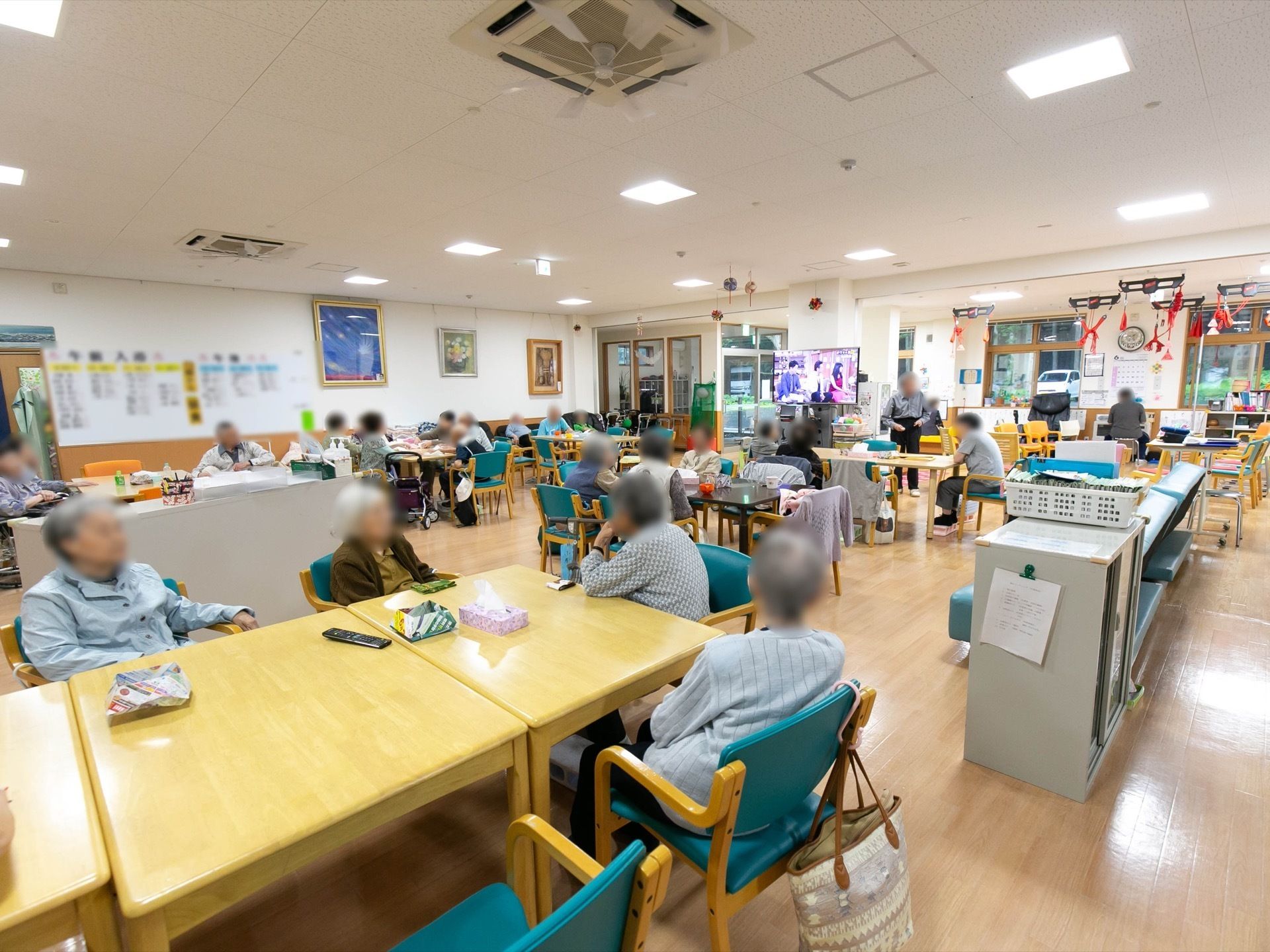 広い食堂に多くの入居者様が集まり、椅子に座ってくつろいでいる。柱に設置されたテレビを見ている方もいる。
