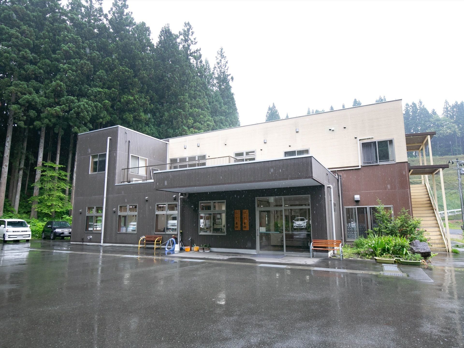 ３色の外壁を持つ２階建ての施設で、玄関前には大きなひさしがある。建物の横には大きな杉が何本も生えている。