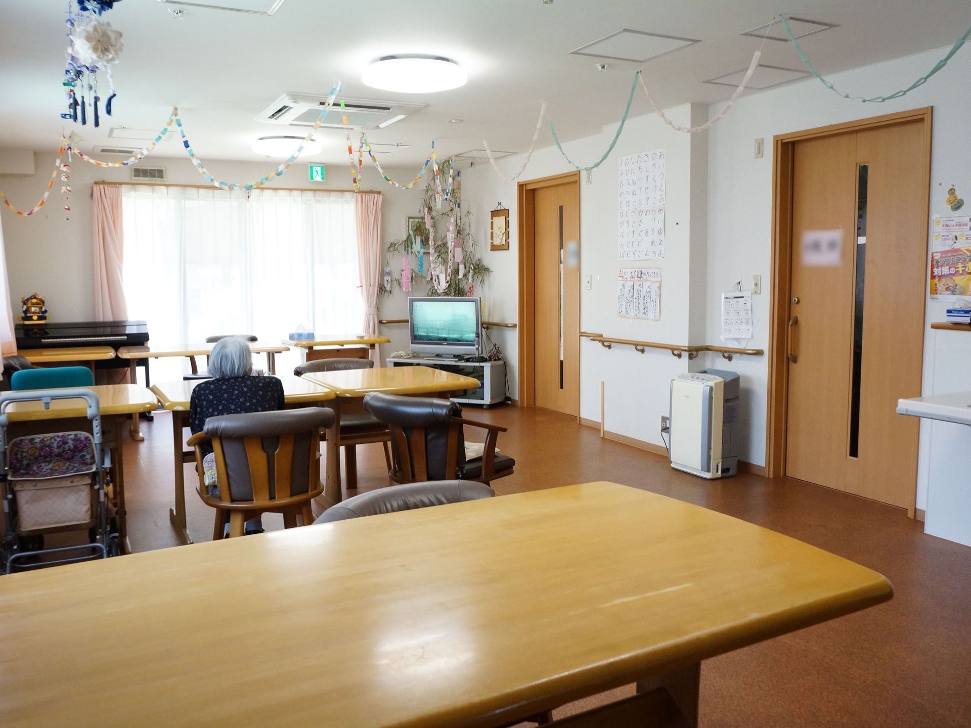 木のテーブルと座り心地の良い椅子が並んだ食堂。天井からは折り紙で作った飾りが吊り下げられており、テレビが置かれている。