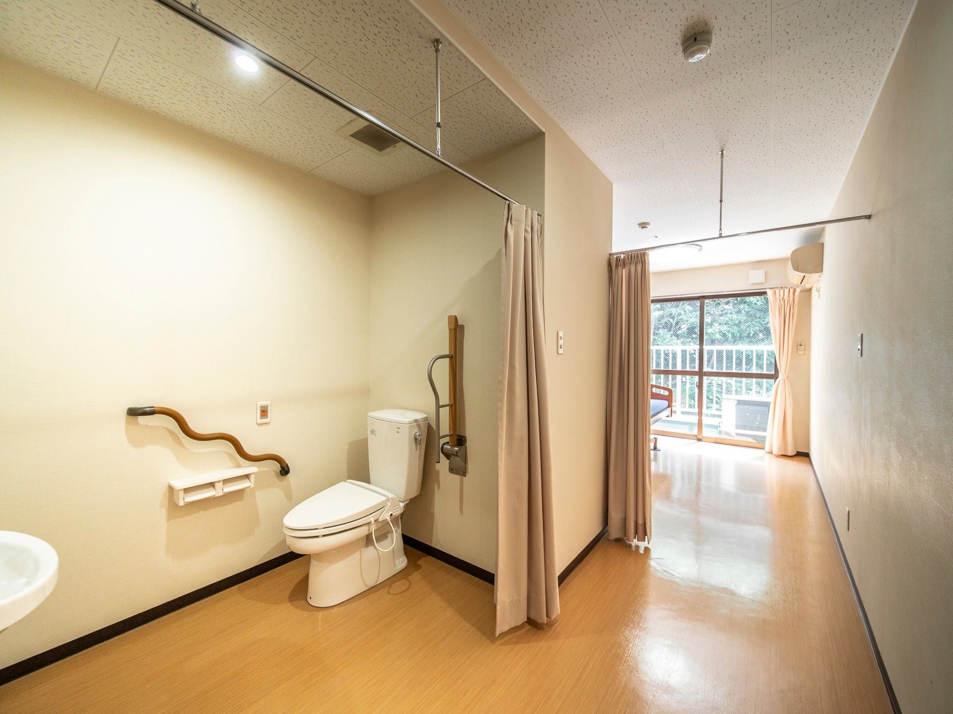 居室にある大きなトイレ。扉はなく、カーテンが掛かっている。壁には手すりと可動式のひじ掛けが付いている。